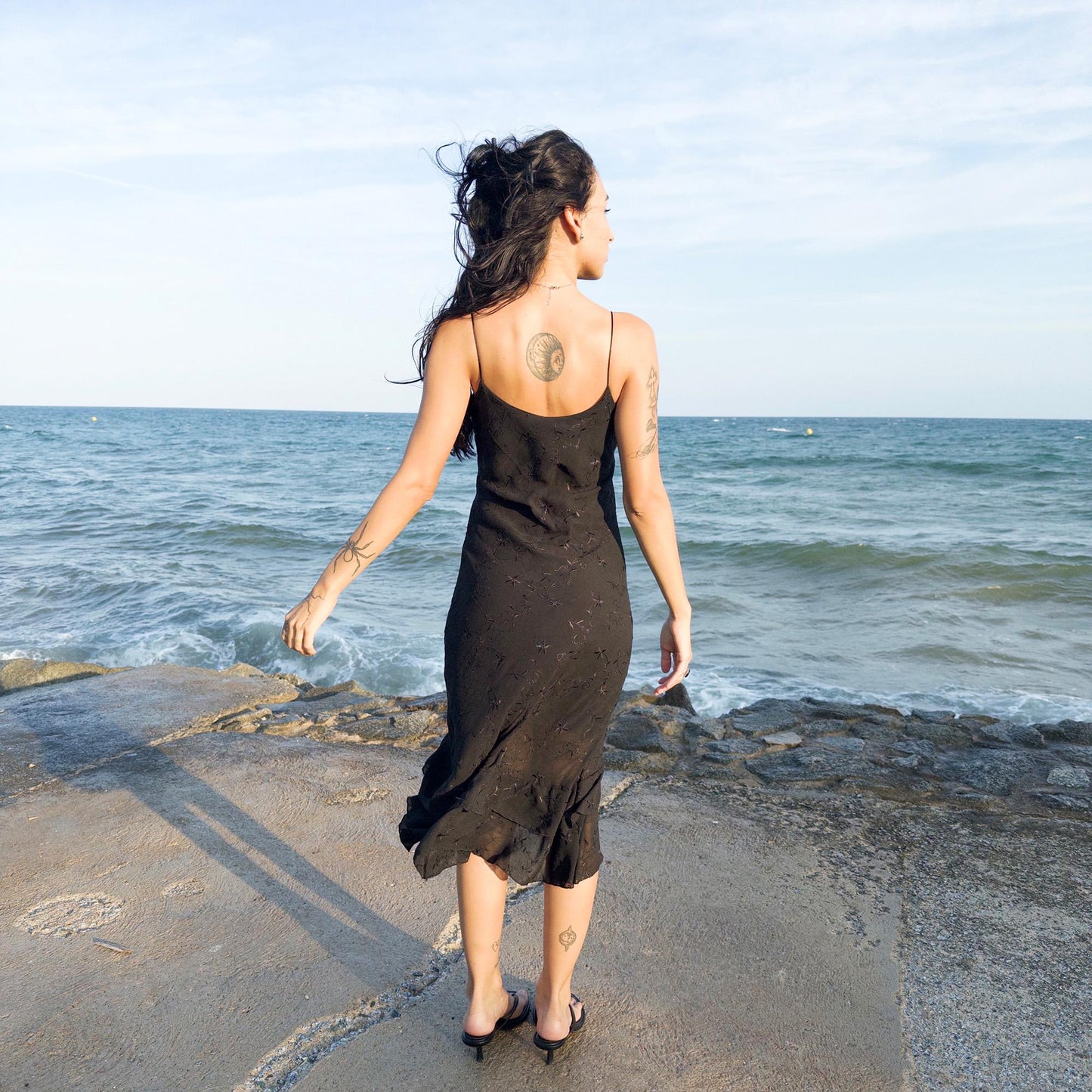 The vintage 2000s black midi dress is a French-made slip silhouette with delicate spaghetti straps and a tiered ruffle hem that drapes asymmetrically. Lightweight with subtle floral embroidery, it flows with ease—ideal for summer promenades, cocktail sunsets, or barefoot evenings. Its understated sensuality and effortless cut evoke a romantic calm. Excellent vintage condition.