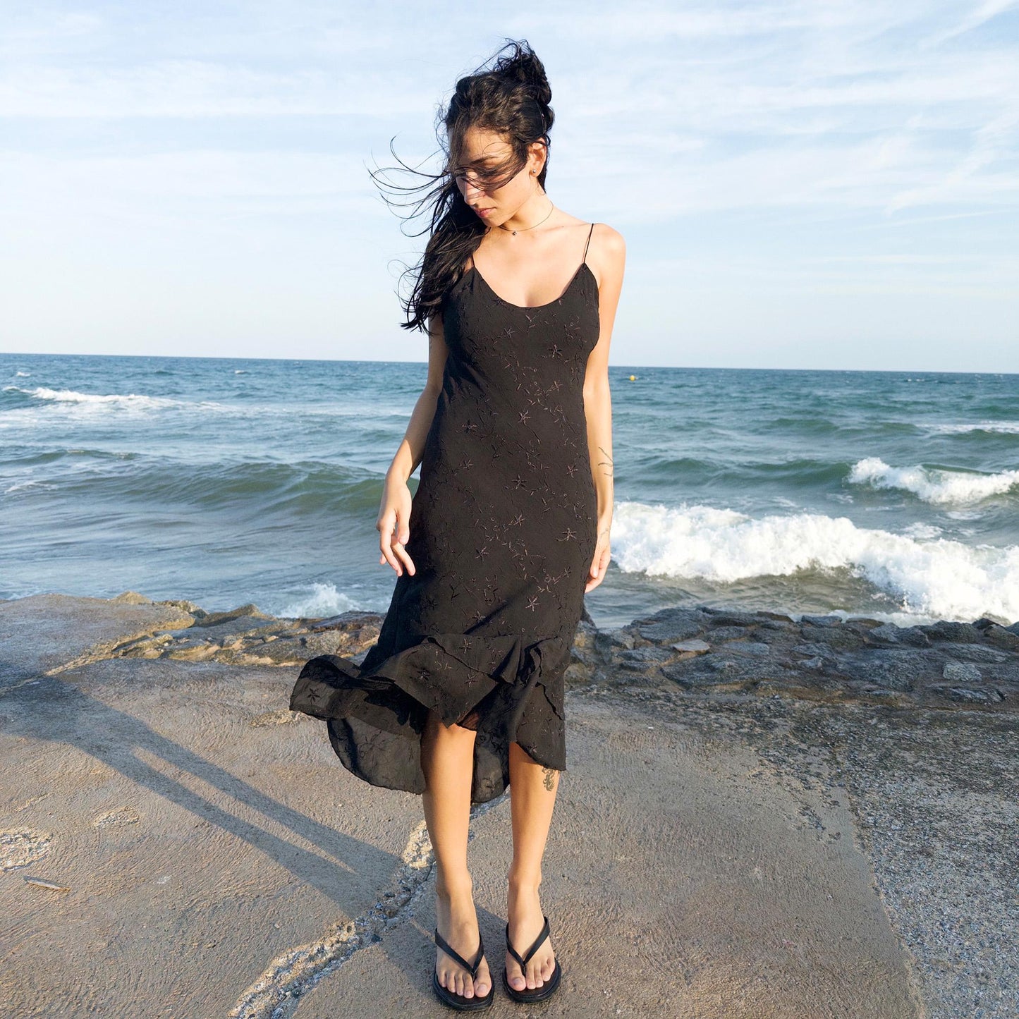 The vintage 2000s black midi dress is a French-made slip silhouette with delicate spaghetti straps and a tiered ruffle hem that drapes asymmetrically. Lightweight with subtle floral embroidery, it flows with ease—ideal for summer promenades, cocktail sunsets, or barefoot evenings. Its understated sensuality and effortless cut evoke a romantic calm. Excellent vintage condition.