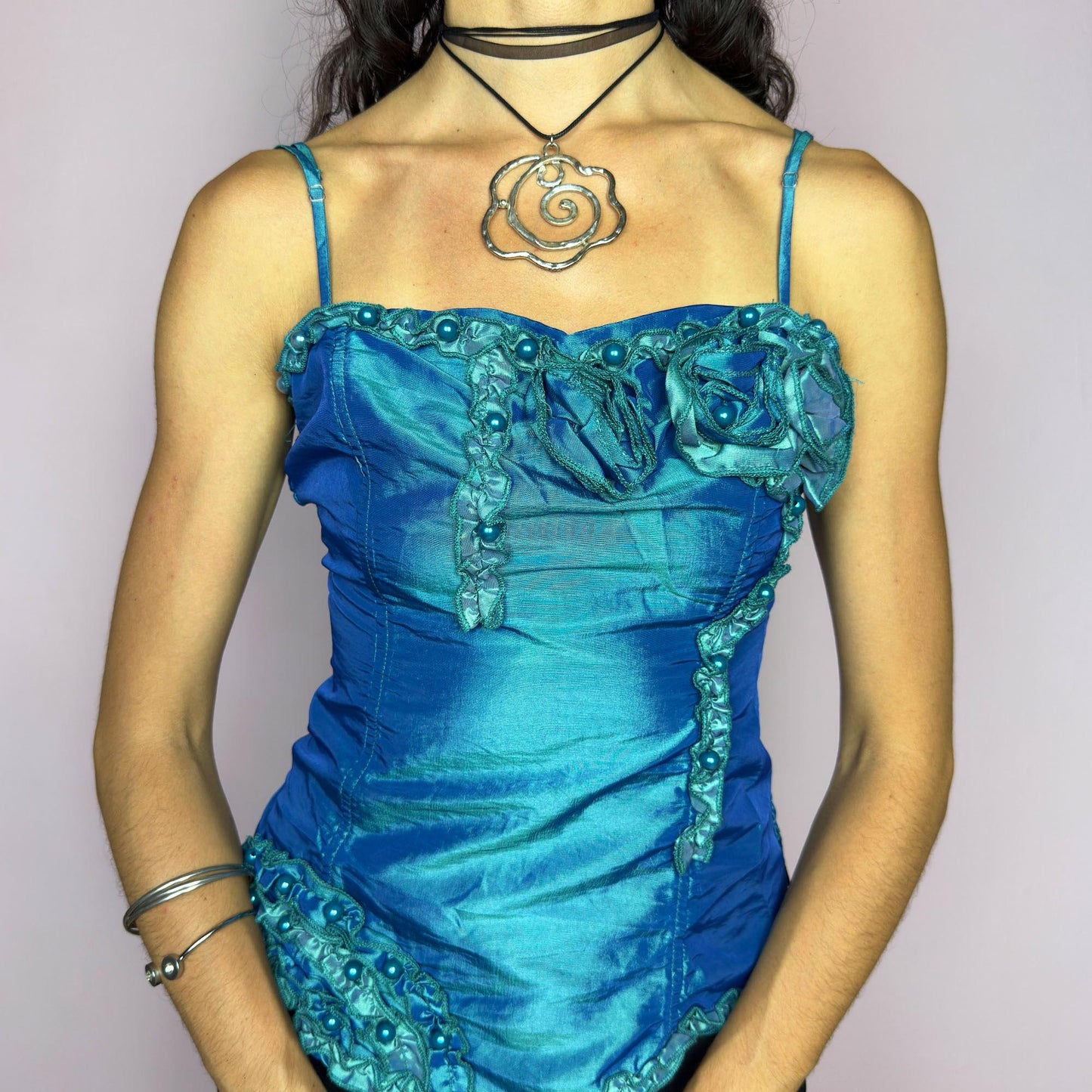 This vintage 2000s iridescent turquoise corset top features adjustable spaghetti straps, floral rosette appliqué, and beaded trim. The structured front sculpts the waist, while the ruched smocked back adds stretch and comfort. An asymmetrical hem and glossy finish elevate it for prom, weddings, or dreamy nights out. A romantic Y2K formal top that blends fairytale opulence with a bold, sculptural silhouette. Excellent vintage condition.