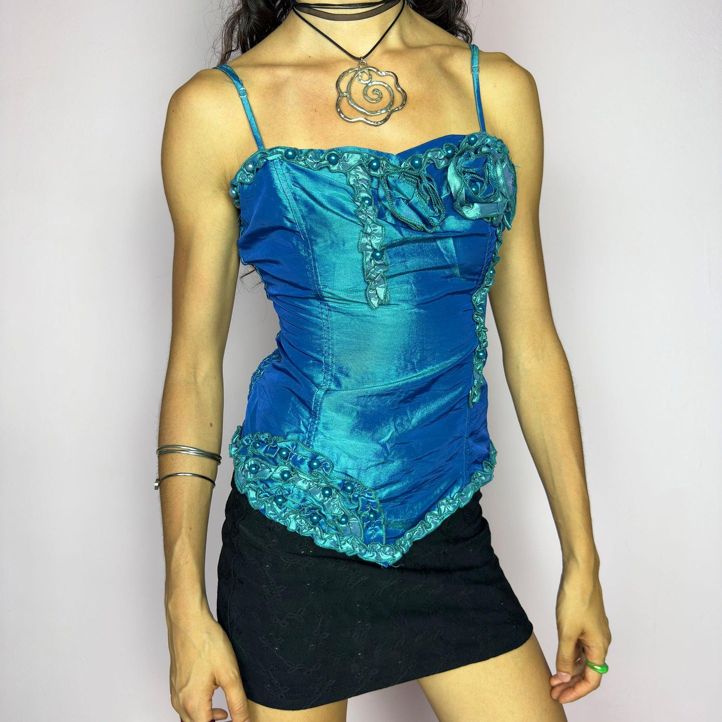 This vintage 2000s iridescent turquoise corset top features adjustable spaghetti straps, floral rosette appliqué, and beaded trim. The structured front sculpts the waist, while the ruched smocked back adds stretch and comfort. An asymmetrical hem and glossy finish elevate it for prom, weddings, or dreamy nights out. A romantic Y2K formal top that blends fairytale opulence with a bold, sculptural silhouette. Excellent vintage condition.