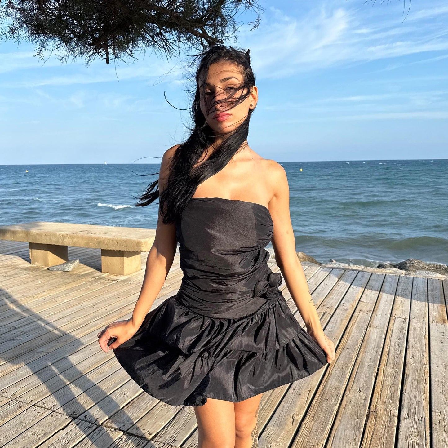 The vintage 90s black mini dress is a sculptural, strapless piece with ruched draping and voluminous double ruffle tiers. Made in Italy, it features a tie-back sash, shirred elastic panel, and side zip for a secure, flattering fit. Crafted from a polyester blend, it catches light effortlessly—ideal for summer nights, cocktail events, or rooftop parties. A super cute dress with high femme drama. Excellent vintage condition.