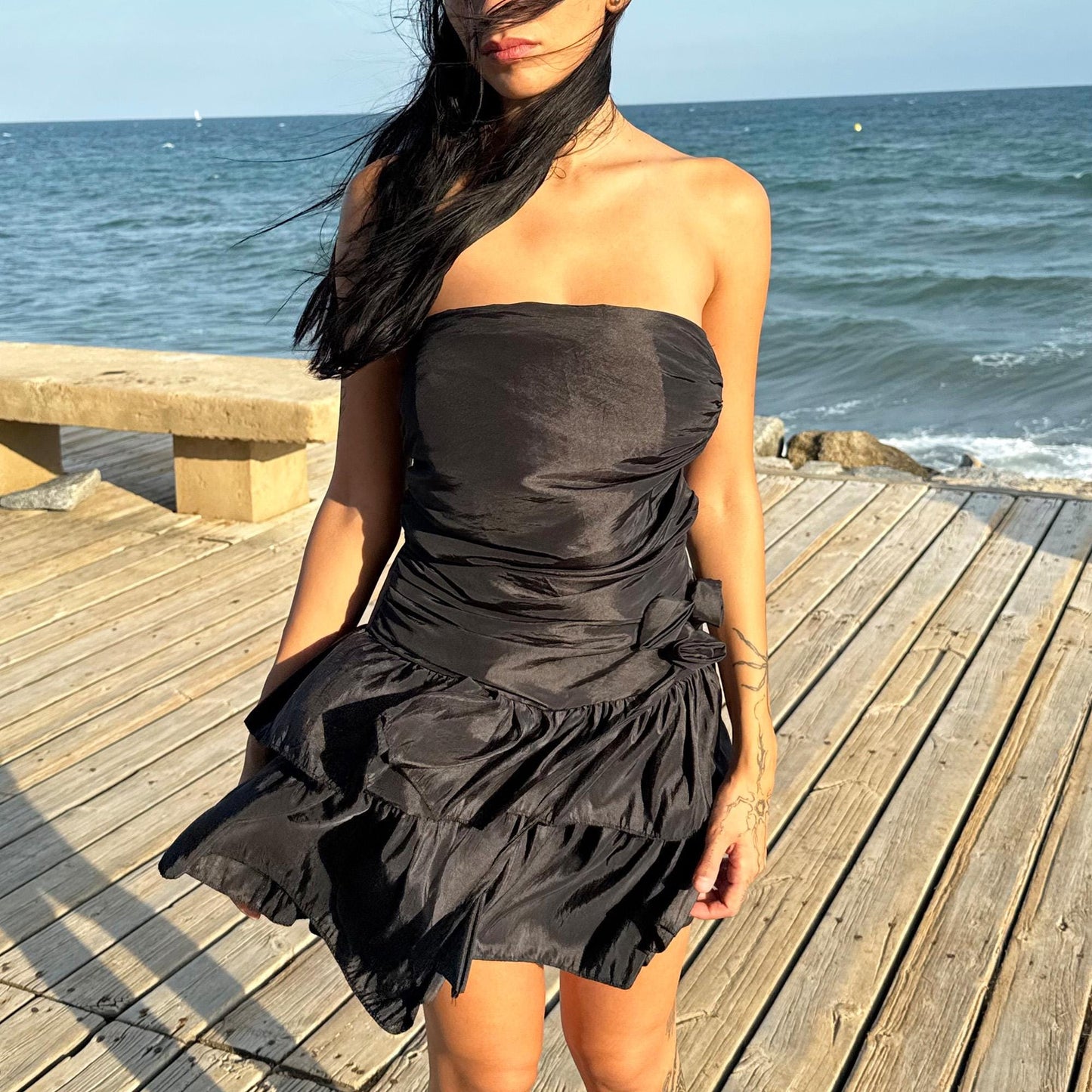 The vintage 90s black mini dress is a sculptural, strapless piece with ruched draping and voluminous double ruffle tiers. Made in Italy, it features a tie-back sash, shirred elastic panel, and side zip for a secure, flattering fit. Crafted from a polyester blend, it catches light effortlessly—ideal for summer nights, cocktail events, or rooftop parties. A super cute dress with high femme drama. Excellent vintage condition.