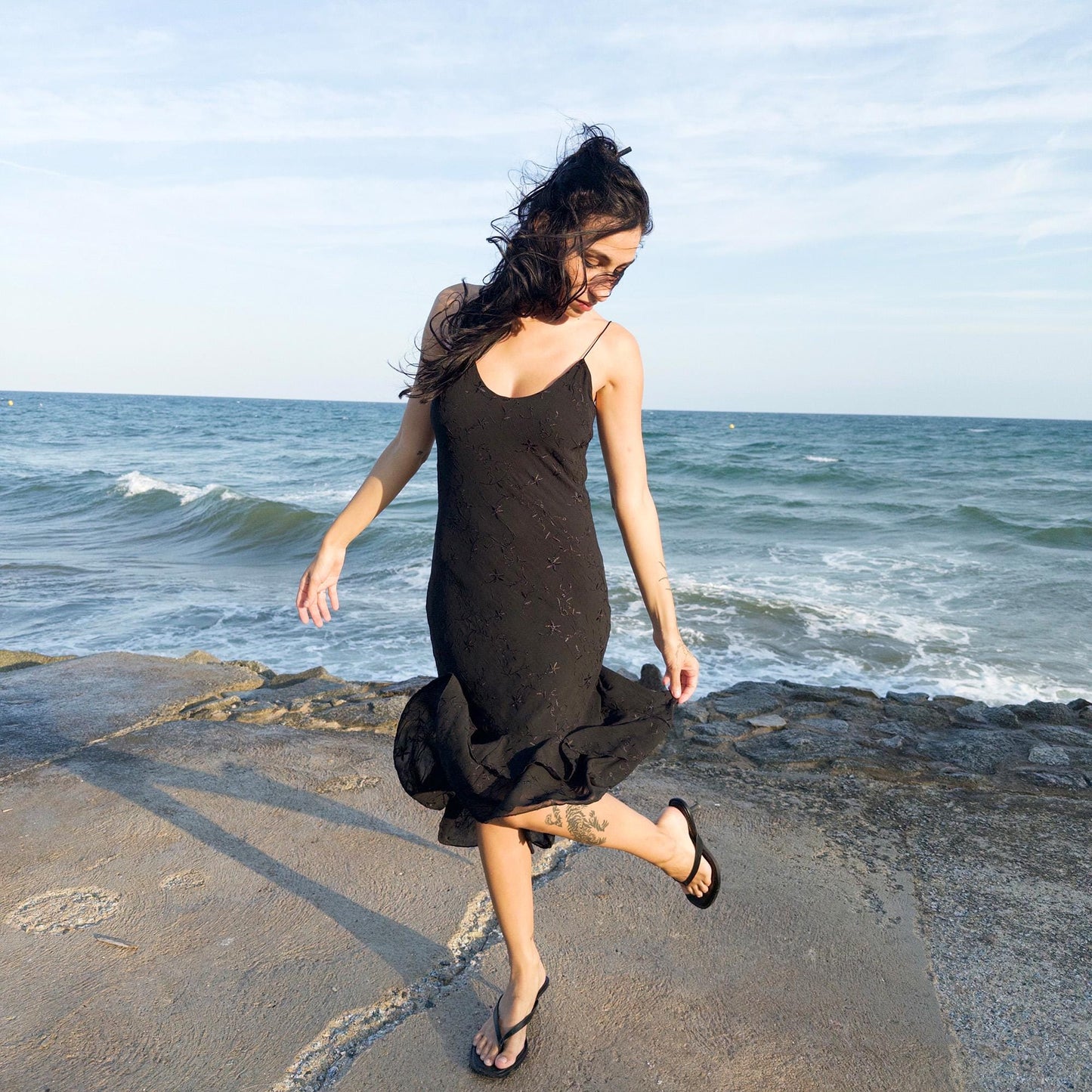 The vintage 2000s black midi dress is a French-made slip silhouette with delicate spaghetti straps and a tiered ruffle hem that drapes asymmetrically. Lightweight with subtle floral embroidery, it flows with ease—ideal for summer promenades, cocktail sunsets, or barefoot evenings. Its understated sensuality and effortless cut evoke a romantic calm. Excellent vintage condition.