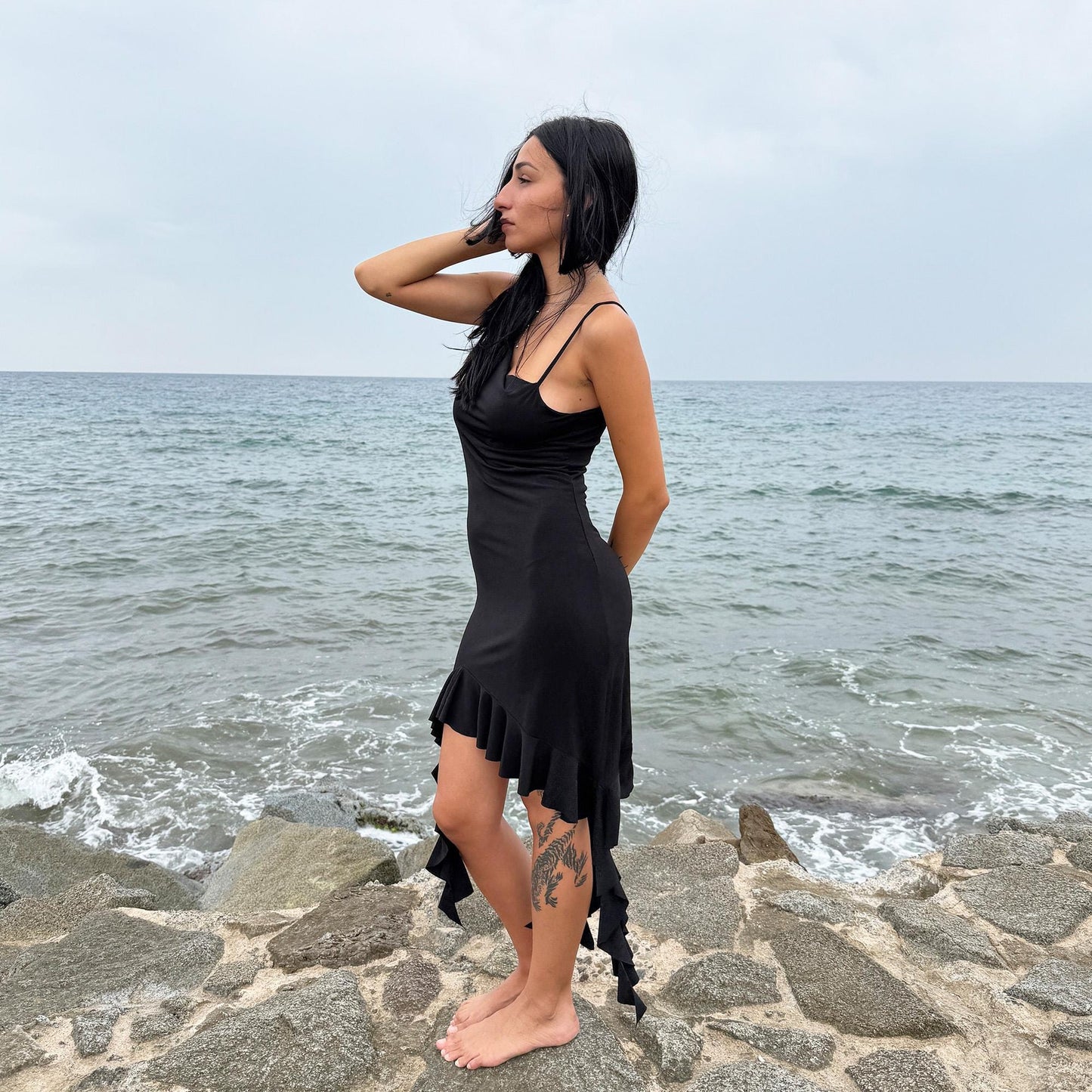 This vintage 2000s black mini dress features asymmetric straps with a subtle cowl neckline and a dramatic ruffled hem. Crafted from a stretchy fabric, it flatters the body with a bold party silhouette. Perfect for summer nights, club looks, or diva-inspired Y2K styling, this piece captures iconic 2000s glamour. Excellent vintage condition.
