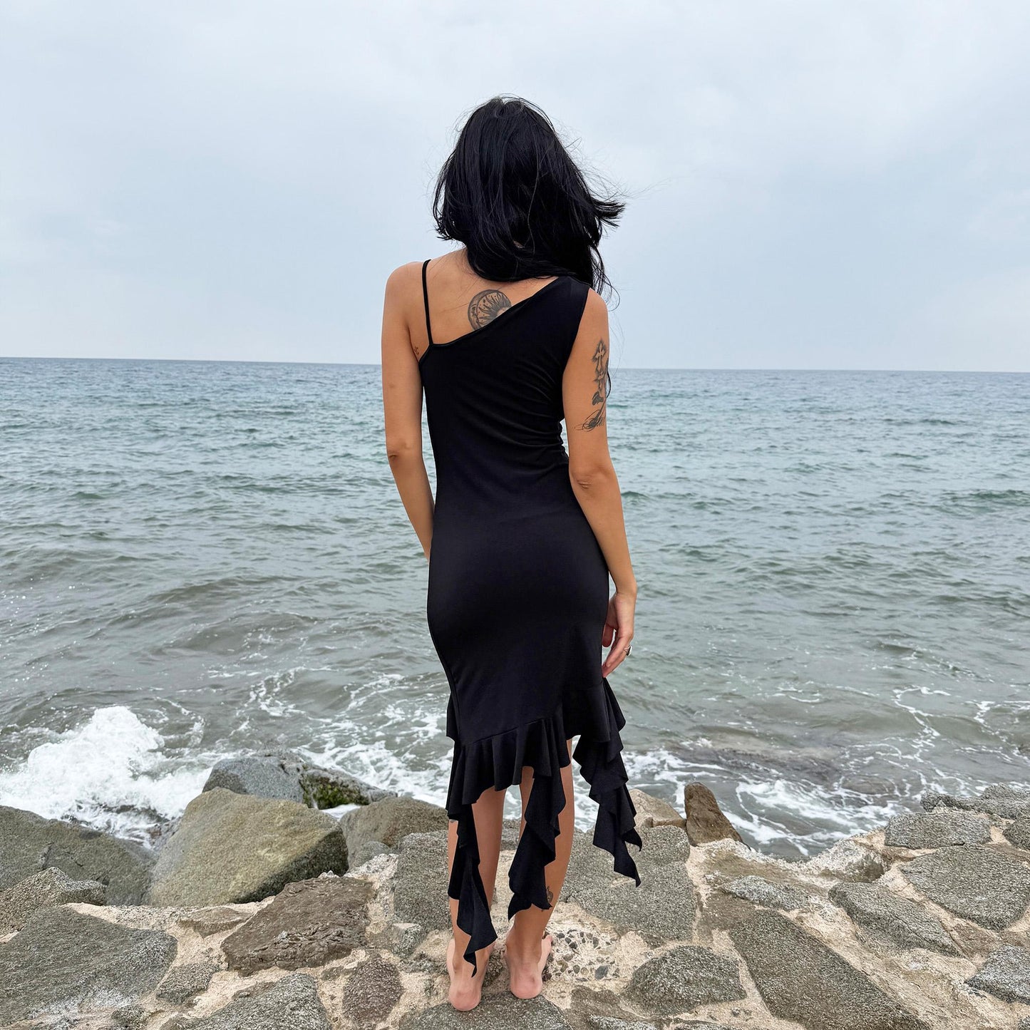 This vintage 2000s black mini dress features asymmetric straps with a subtle cowl neckline and a dramatic ruffled hem. Crafted from a stretchy fabric, it flatters the body with a bold party silhouette. Perfect for summer nights, club looks, or diva-inspired Y2K styling, this piece captures iconic 2000s glamour. Excellent vintage condition.