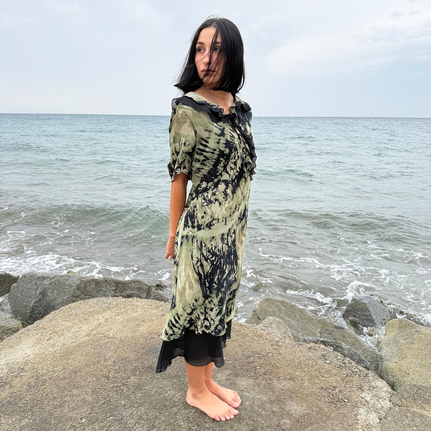 This vintage 90s midi dress features a black and green tie-dye pattern with a ruffled neckline. The short sleeves and flowy layered hem make it ideal for summer festivals, boho gatherings, or dark fairy-inspired looks. A unique Italian-made piece, perfect for forest parties or artistic styling. Made in Italy from lightweight non-stretch fabric. Excellent vintage condition.