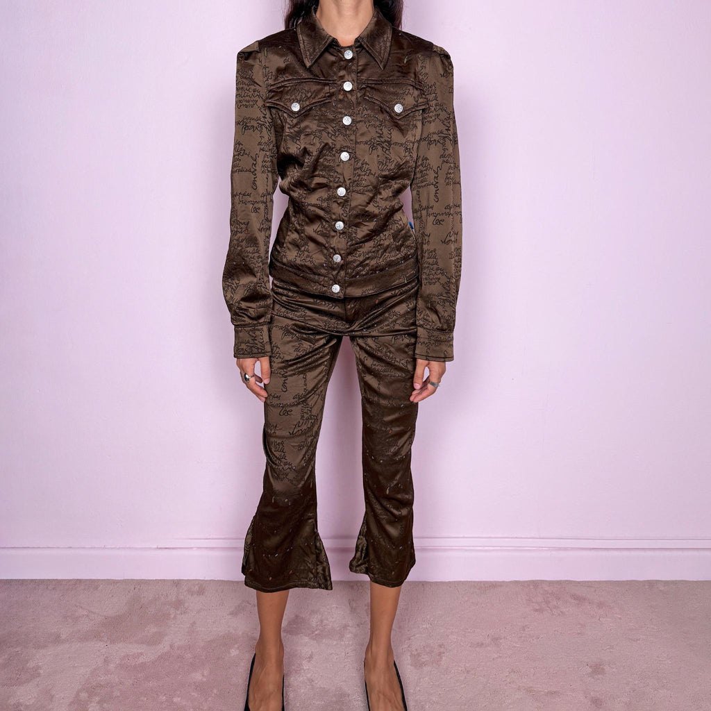 Vintage late 1990s two-piece set from French fashion house Christian Lacroix, from the Jeans de Christian Lacroix line, made in Italy. The set features a brown printed jacket with a glossy, satin-like finish, handwritten-style graphic motif, pointed collar, flap chest pockets, and branded snap-button closure, paired with matching high-waisted cropped trousers with a slim fit through the leg and flared hems. Crafted from a stretchy blend of acetate, polyamide, polyester, and elastane.