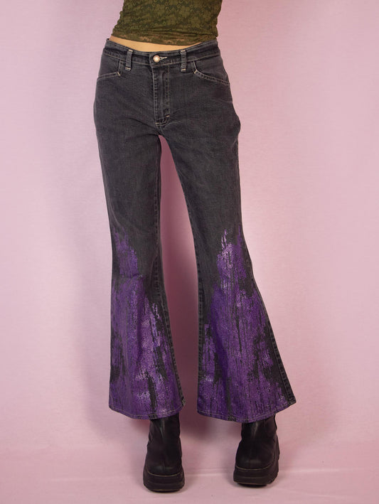 The Y2K Gray Flare Jeans are a vintage 2000s mid-rise pants featuring stretchy gray denim with purple glitter paint splatter detailing on the flared legs, front pockets, and a zipper closure. Perfect for festival looks or casual outfits. Made in UK.