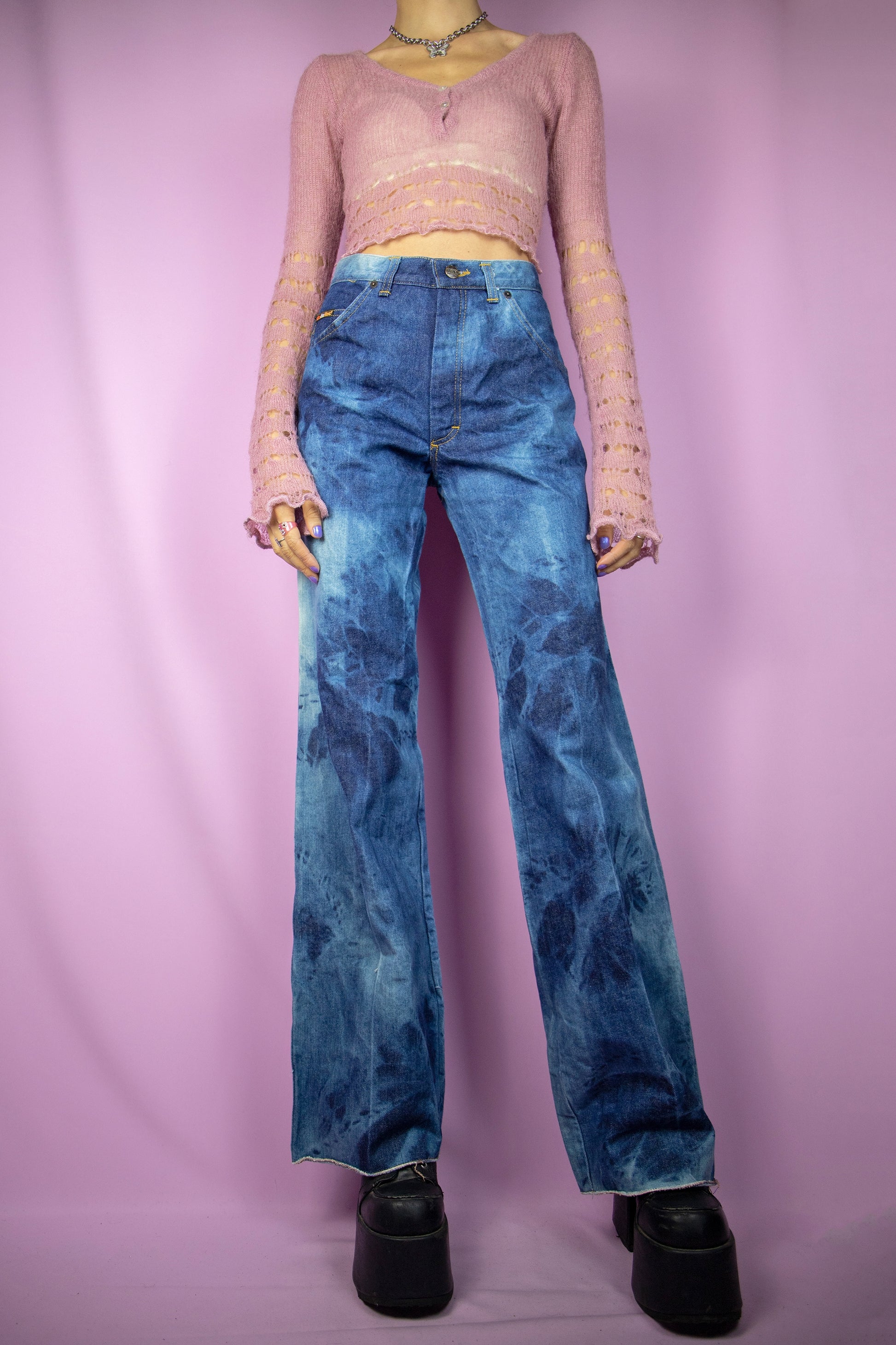 The Vintage 90s Tie Dye Wide Jeans feature a high-waisted design with a relaxed, wide-leg silhouette. These jeans showcase a bold tie-dye wash in varying shades of blue and are finished with a zipper closure, pockets, and a raw hem. Perfect for festivals and casual wear.