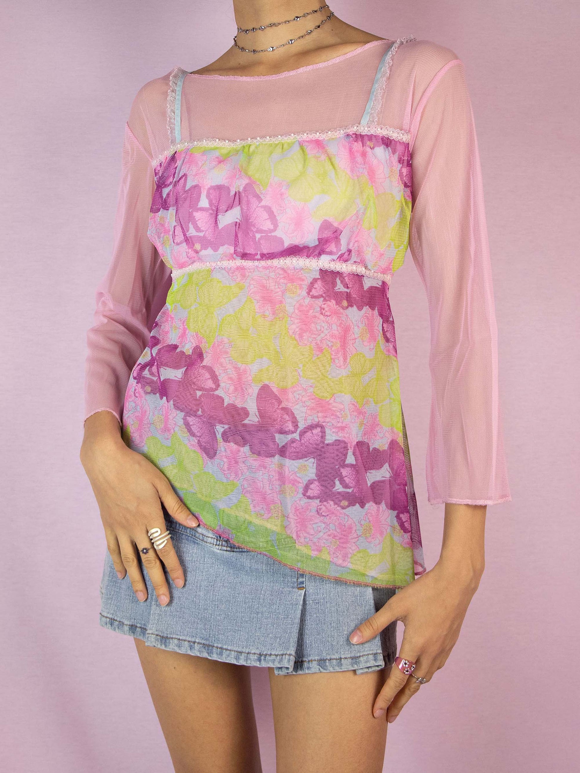 The Y2K Pink Printed Mesh Top is a vintage 2000s romantic semi-sheer top in light pink and green, adorned with whimsical floral and butterfly graphics. Featuring three-quarter sleeves and an asymmetric hem, this enchanting piece adds a fairy-like and playful touch to any party or special event ensemble. Excellent vintage condition.