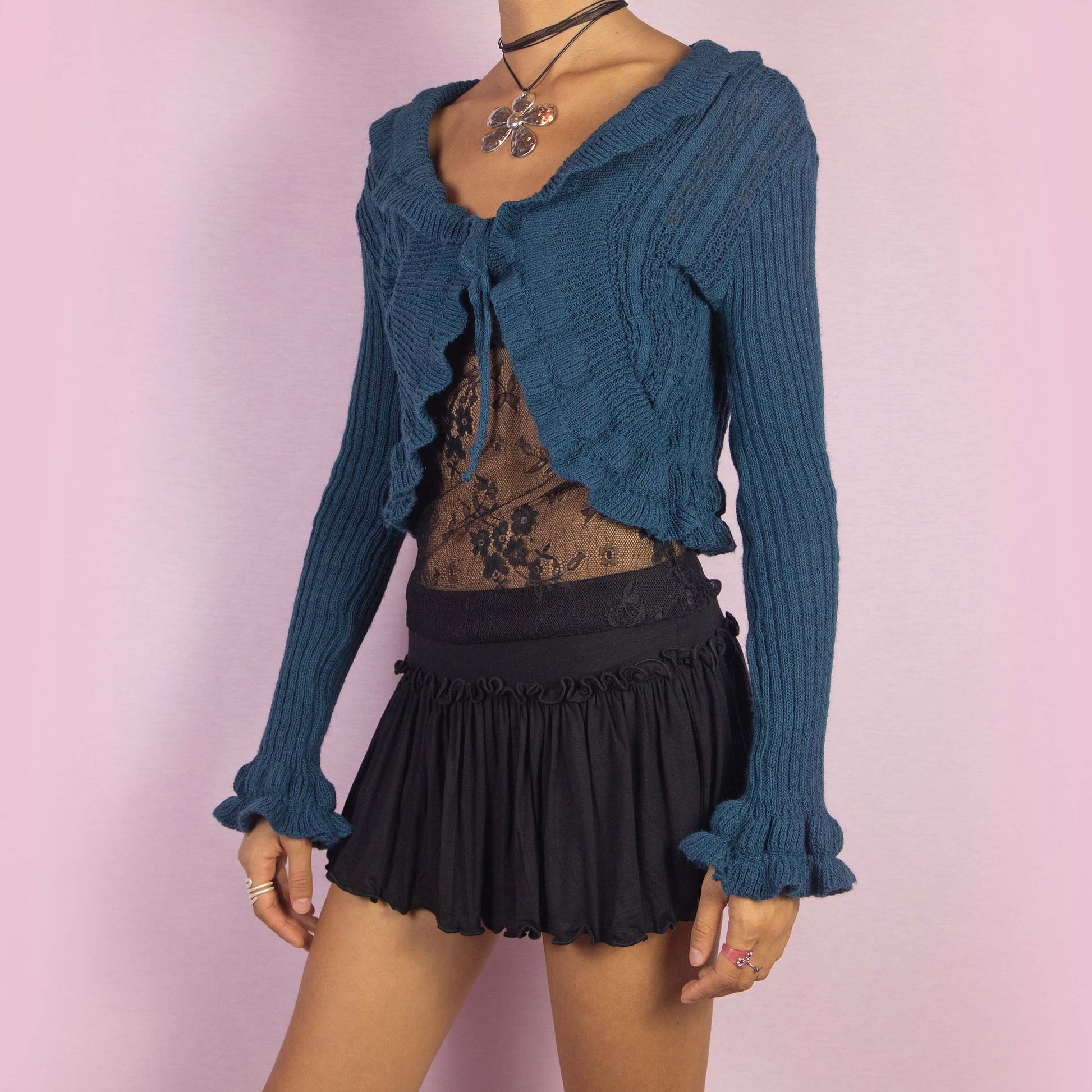 The Y2K Blue Ruffle Cropped Cardigan is a vintage 2000s romantic bolero crafted from a cozy wool blend, featuring a flattering tie-front design and playful ruffle details along the hem, sleeves, and neckline. Perfect for autumn or cooler spring days.