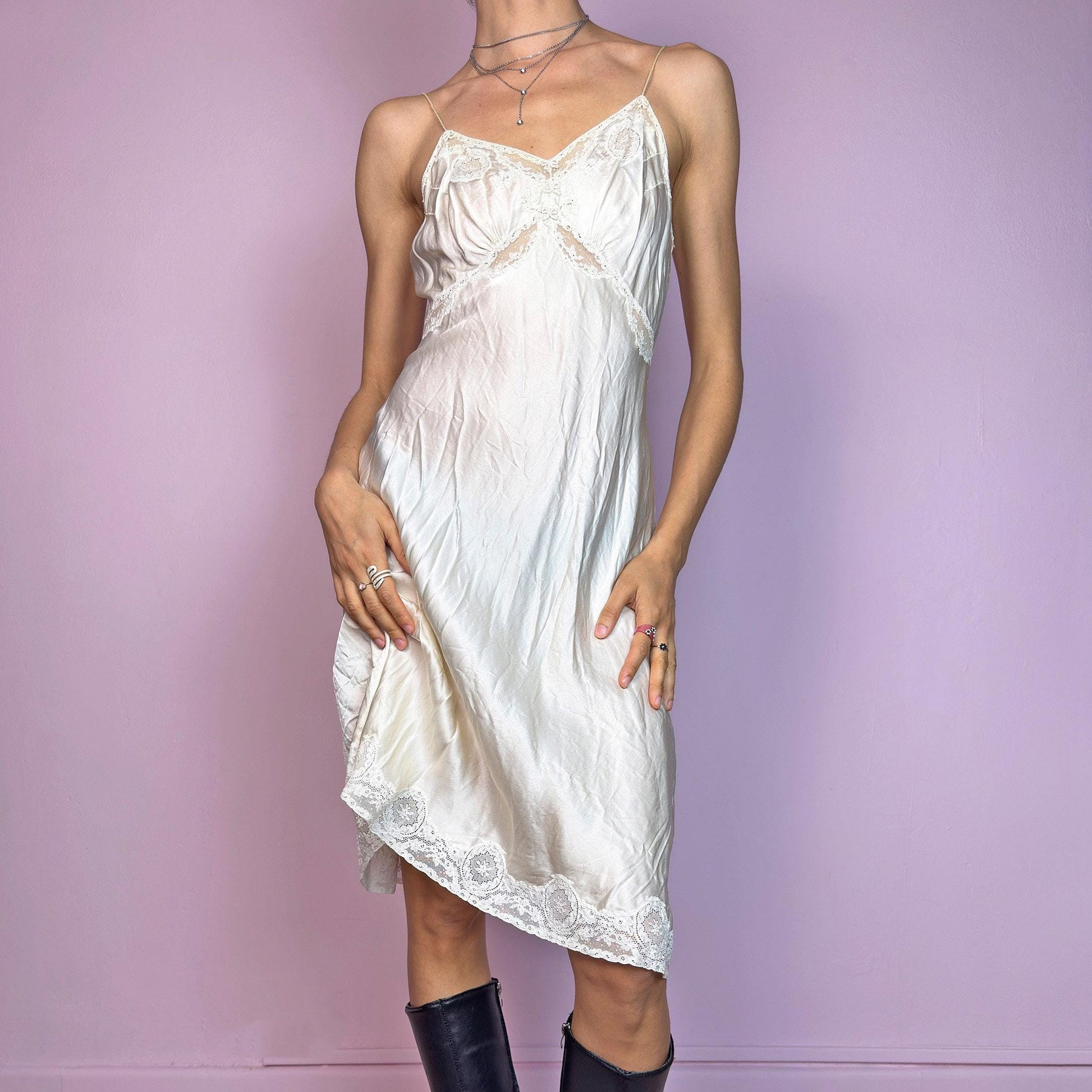 This vintage cream midi slip dress is likely from the 1940s–1950s, crafted from satin-style fabric with intricate floral lace at the bust, waist, and hem. Features a soft V-neckline, thin non-adjustable straps, and a body-skimming bias cut for graceful movement. Falls below the knee with a fluid, vintage silhouette. A rare, romantic undergarment with timeless elegance—ideal for collectors, layering, or as an ethereal evening statement.