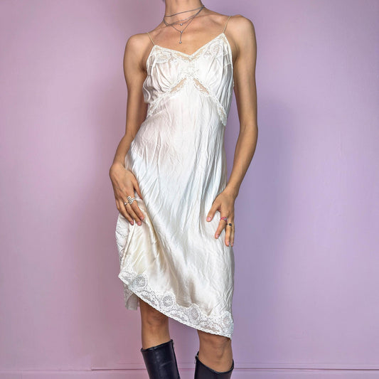 This vintage cream midi slip dress is likely from the 1940s–1950s, crafted from satin-style fabric with intricate floral lace at the bust, waist, and hem. Features a soft V-neckline, thin non-adjustable straps, and a body-skimming bias cut for graceful movement. Falls below the knee with a fluid, vintage silhouette. A rare, romantic undergarment with timeless elegance—ideal for collectors, layering, or as an ethereal evening statement.