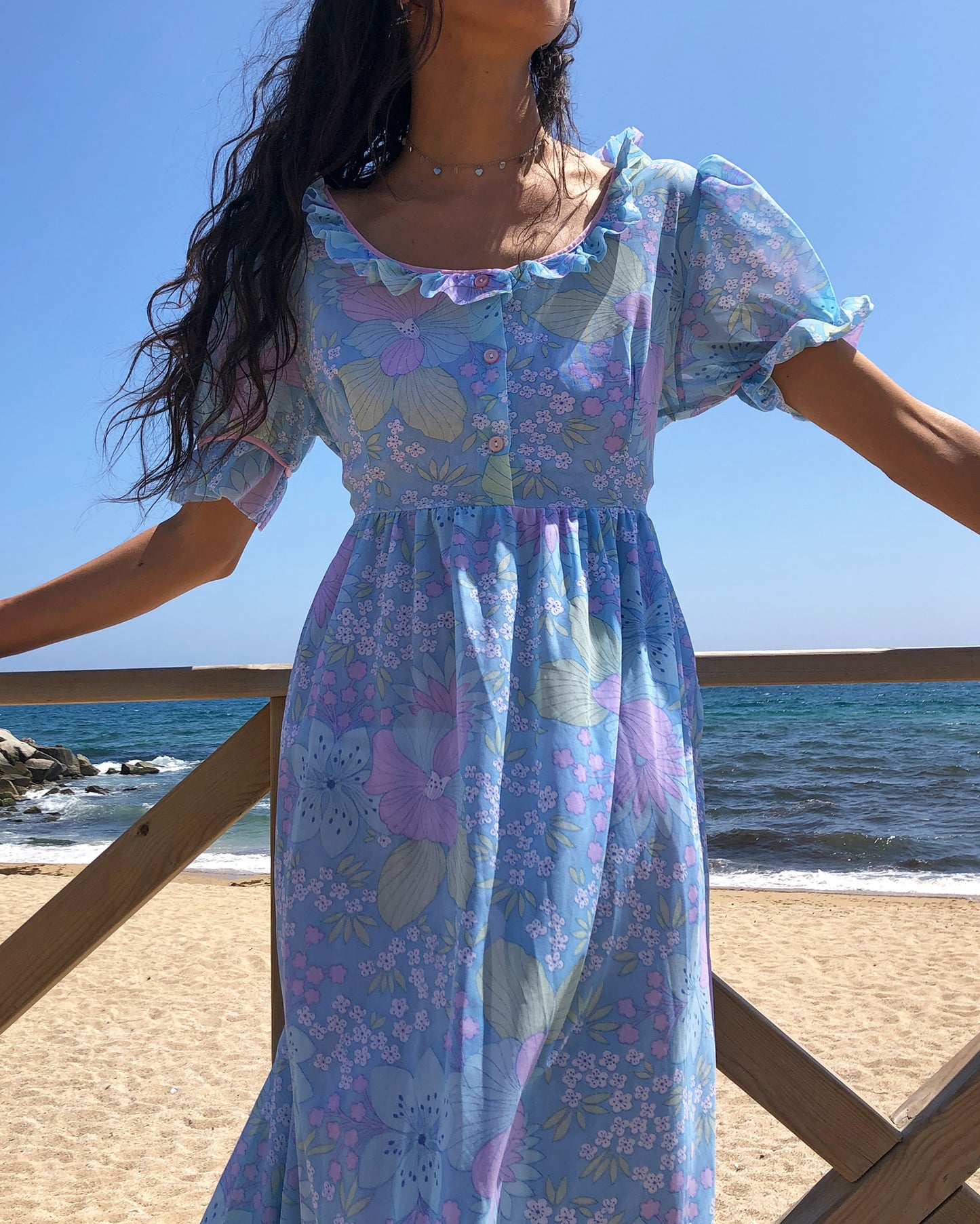This vintage 1970s blue floral midi dress features lightweight semi-sheer fabric in soft pastel tones with a square neckline and ruffle trim, puff sleeves with frilled cuffs, and a button-front bodice. The empire waist and flowing length create gentle prairie romance for spring picnics, cottagecore styling, or feminine nostalgia. Made in France from a polyamide nylon blend. Excellent vintage condition.