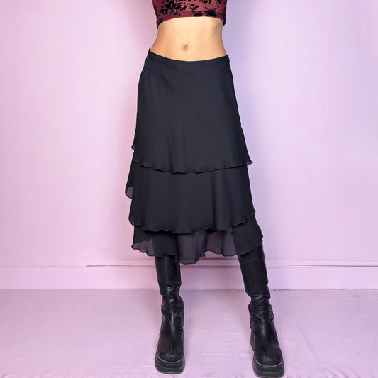 The Vintage 90s Black Layered Tiered Skirt features a flowy, tiered design with cascading chiffon-like layers. It includes a side zipper closure. Its versatile length and elegant style make it perfect for evening parties. Adult World Shop