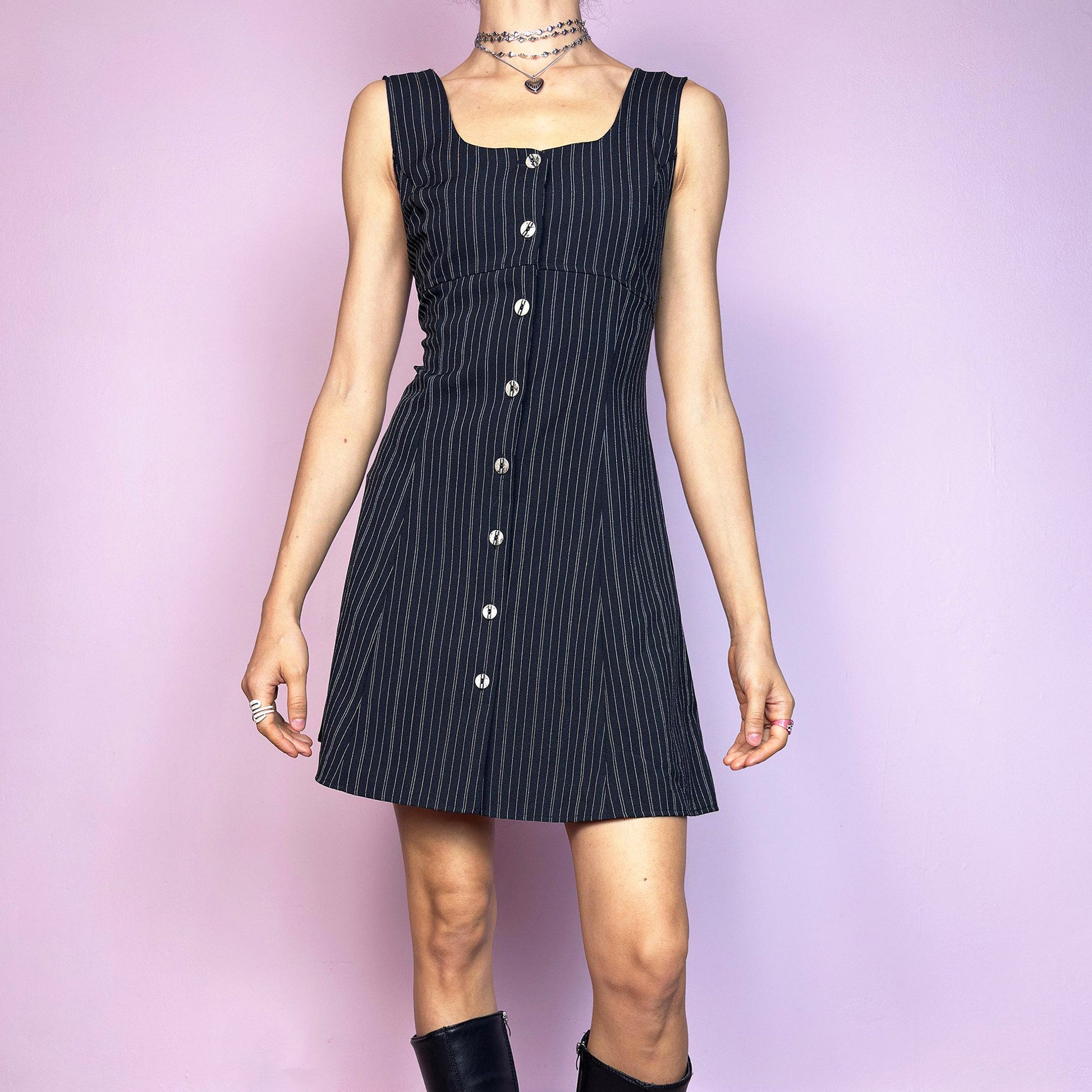 The Vintage 90s Black Pinstripe Mini Dress is a timeless piece with a flirty twist. This sleeveless dress features a black base with fine pinstripes, a fitted bodice with a square neckline, and a full front button closure. The slight A-line silhouette flatters the waist while keeping the vibe playful and polished. Ideal for 90s office-core and preppy looks. Excellent vintage condition.