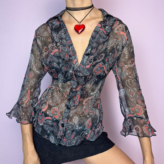 The Vintage 90s Black Ruffle Sheer Blouse features a semi-sheer fabric with an allover paisley print in red, grey, and white tones. This fitted silhouette includes three-quarter bell sleeves with flared ruffle cuffs, a romantic ruffled collar, and a front button-up closure. The lightweight fabric makes it perfect for summer layering or wearing on its own for a sleek, sexy vibe. Great vintage condition. adult world shop