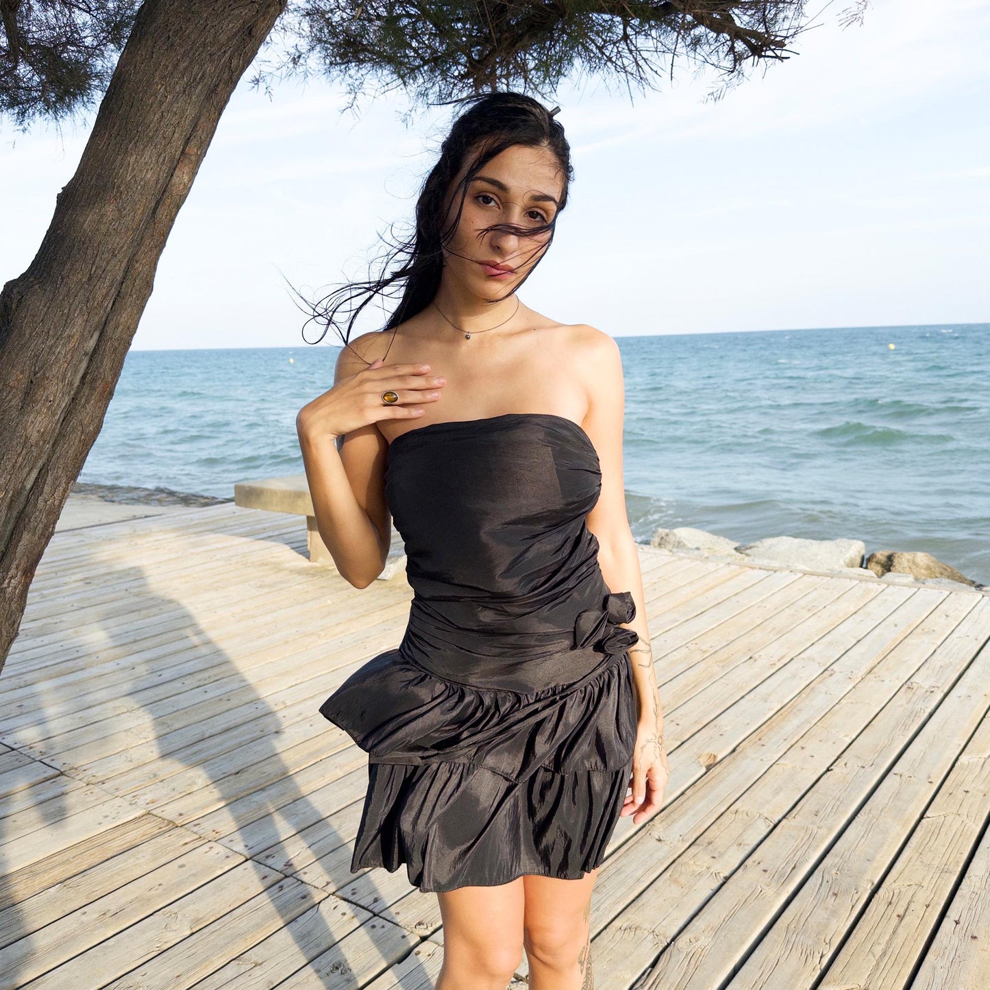 The vintage 90s black mini dress is a sculptural, strapless piece with ruched draping and voluminous double ruffle tiers. Made in Italy, it features a tie-back sash, shirred elastic panel, and side zip for a secure, flattering fit. Crafted from a polyester blend, it catches light effortlessly—ideal for summer nights, cocktail events, or rooftop parties. A super cute dress with high femme drama. Excellent vintage condition.