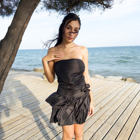 The vintage 90s black mini dress is a sculptural, strapless piece with ruched draping and voluminous double ruffle tiers. Made in Italy, it features a tie-back sash, shirred elastic panel, and side zip for a secure, flattering fit. Crafted from a polyester blend, it catches light effortlessly—ideal for summer nights, cocktail events, or rooftop parties. A super cute dress with high femme drama. Excellent vintage condition.