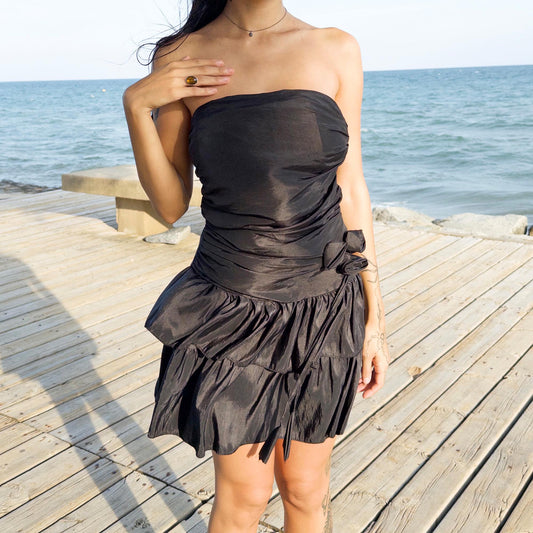 The vintage 90s black mini dress is a sculptural, strapless piece with ruched draping and voluminous double ruffle tiers. Made in Italy, it features a tie-back sash, shirred elastic panel, and side zip for a secure, flattering fit. Crafted from a polyester blend, it catches light effortlessly—ideal for summer nights, cocktail events, or rooftop parties. A super cute dress with high femme drama. Excellent vintage condition.