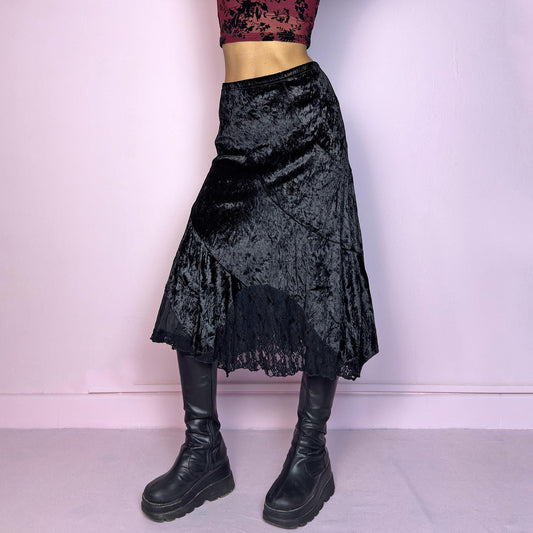 The Vintage 90s Black Velvet Asymmetric Skirt features a velvet fabric with an elegant asymmetrical hemline adorned with lace detailing for a touch of whimsy and an elastic waist. Its romantic and gothic-inspired design. Adult World Shop
