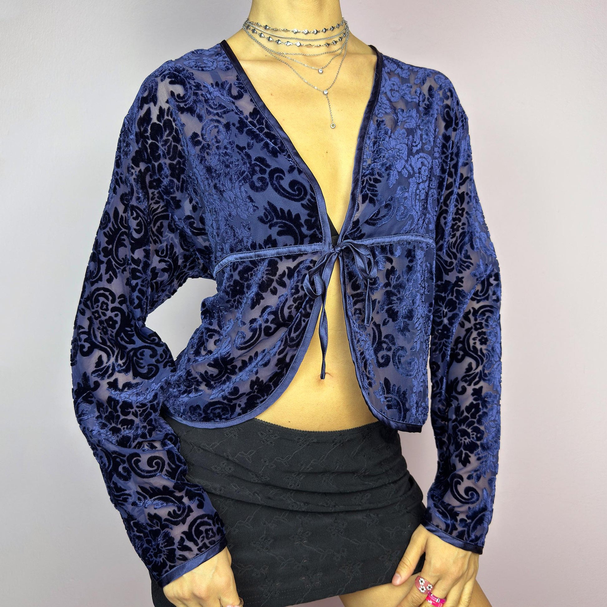 The Vintage 90s Blue Velvet Sheer Blouse is a romantic sheer velvet top featuring a flocked baroque floral pattern on deep navy blue. Designed with a plunging V-neckline, long sleeves, and a delicate tie-front closure, this blouse is perfect for night-out looks. adult world shop