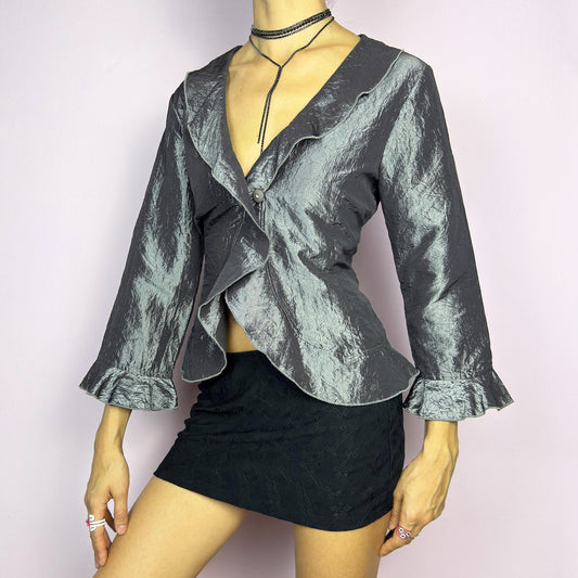 The Vintage 90s Gray Iridescent Ruffle Blouse is a sophisticated and elegant piece with a shimmering iridescent finish in a metallic gray tone. It features a crinkled texture, a cascading ruffle trims along the hemline and three-quarter sleeves. Designed with a flattering deep neckline and a single-button closure, this blouse is perfect for formal events or evening wear. Great vintage condition.