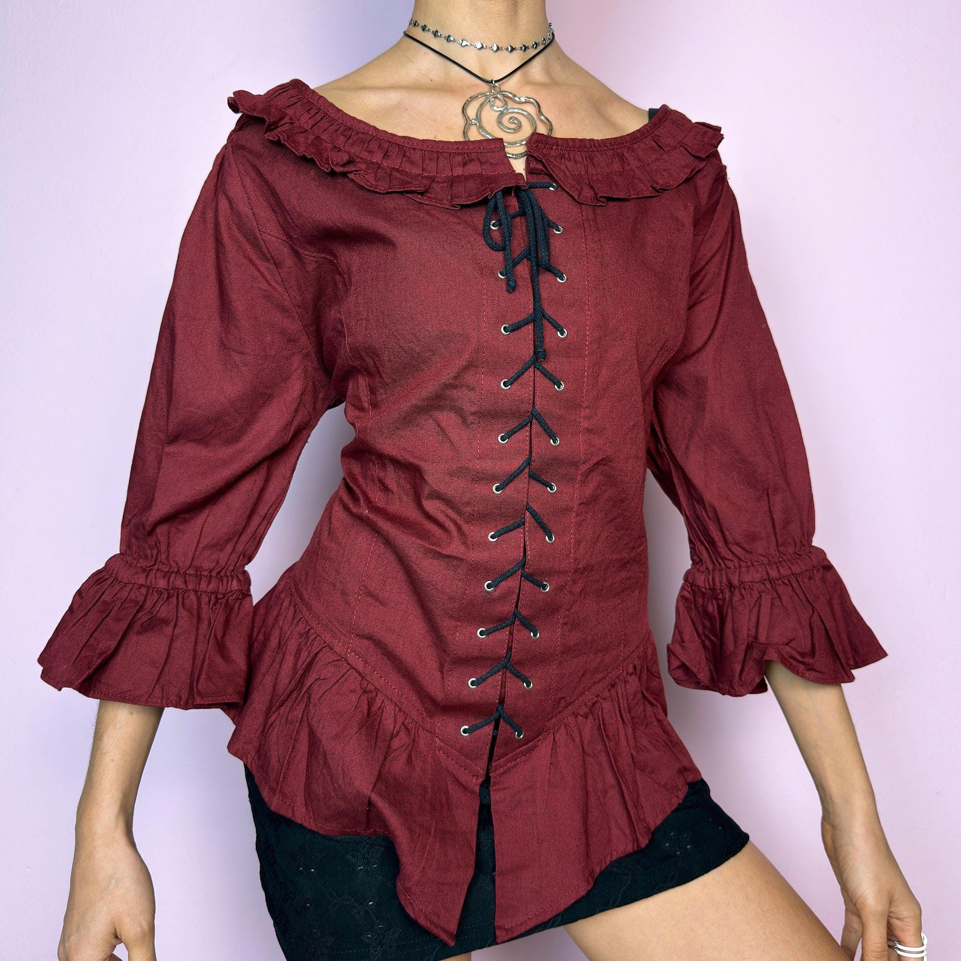 The Vintage 90s Maroon Lace Up Top is a romantic and dramatic piece inspired by gothic and cottagecore aesthetics. Crafted in a deep wine red tone, it features front and back corset-style lace-up details for an adjustable fit. The design includes a ruffled off-the-shoulder neckline, voluminous three-quarter sleeves with gathered ruffle cuffs, and a flattering cinched waist with a ruffled hem. Perfect for creating fairytale-inspired outfits or statement festival looks. adult world shop