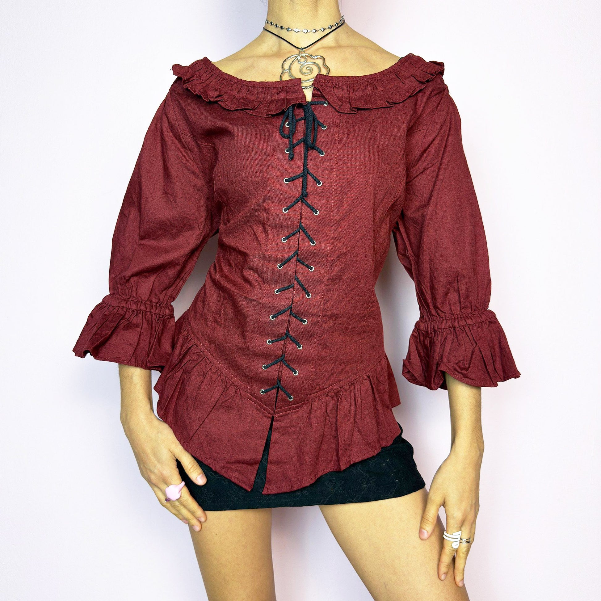 The Vintage 90s Maroon Lace Up Top is a romantic and dramatic piece inspired by gothic and cottagecore aesthetics. Crafted in a deep wine red tone, it features front and back corset-style lace-up details for an adjustable fit. The design includes a ruffled off-the-shoulder neckline, voluminous three-quarter sleeves with gathered ruffle cuffs, and a flattering cinched waist with a ruffled hem. Perfect for creating fairytale-inspired outfits or statement festival looks. Excellent vintage condition.