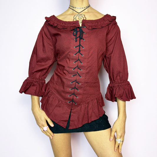 The Vintage 90s Maroon Lace Up Top is a romantic and dramatic piece inspired by gothic and cottagecore aesthetics. Crafted in a deep wine red tone, it features front and back corset-style lace-up details for an adjustable fit. The design includes a ruffled off-the-shoulder neckline, voluminous three-quarter sleeves with gathered ruffle cuffs, and a flattering cinched waist with a ruffled hem. Perfect for creating fairytale-inspired outfits or statement festival looks. Excellent vintage condition.