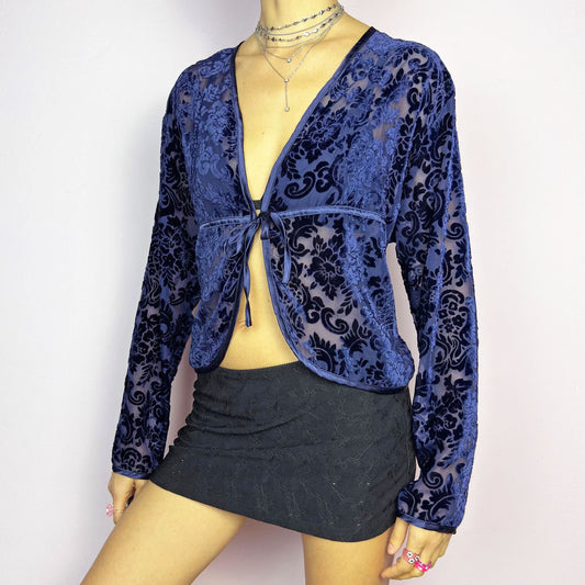 The Vintage 90s Blue Velvet Sheer Blouse is a romantic sheer velvet top featuring a flocked baroque floral pattern on deep navy blue. Designed with a plunging V-neckline, long sleeves, and a delicate tie-front closure, this blouse is perfect for night-out looks.