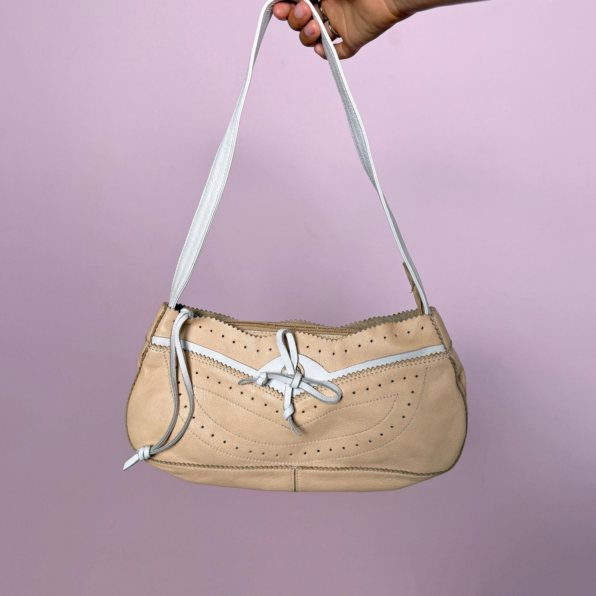 Vintage 2000s shoulder bag, made in Italy from genuine leather in a soft pastel beige and white colorway. Designed in a light baguette-style silhouette, it features a zip-top closure, a white shoulder strap, perforated detailing, contrast stitching, scalloped trim edges, and a small leather tie detail at the front. A delicate and light Y2K piece with a romantic summer feel. From an Italian brand. Excellent vintage condition.