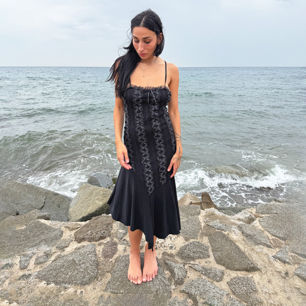 Vintage 2000s black midi dress featuring a fitted silhouette with slim spaghetti straps, delicate ruffled trim along the neckline, vertical ribbon detailing through the bodice, and an asymmetric handkerchief-style hem. Perfect for witchy styling, fairy-inspired looks, and alternative evening outfits. From a French brand, made in France from a stretchy blend of 95% polyester and 5% elastane. Excellent vintage condition.