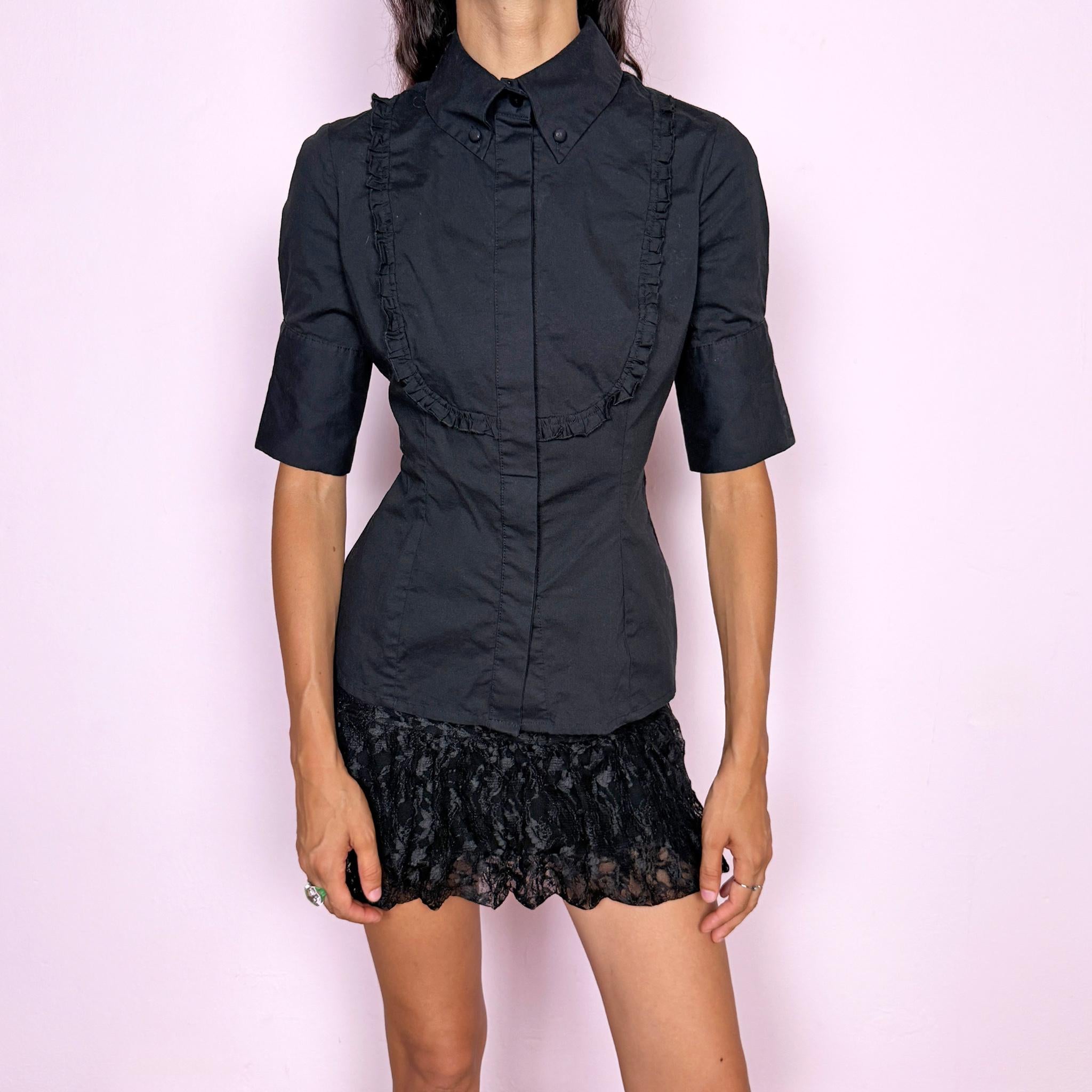 Vintage 2000s black fitted shirt top crafted from a slightly stretchy blend of 97% cotton and 3% elastane. Featuring a structured pointed collar, concealed front button closure, and sculpted panel seams that create a sharply tailored silhouette, finished with ruffled bib detailing across the chest and short sleeves. A refined piece balancing formal tailoring with subtle romantic detailing, ideal for elegant minimalist or dark feminine styling. By Spanish designer Adolfo Domínguez.