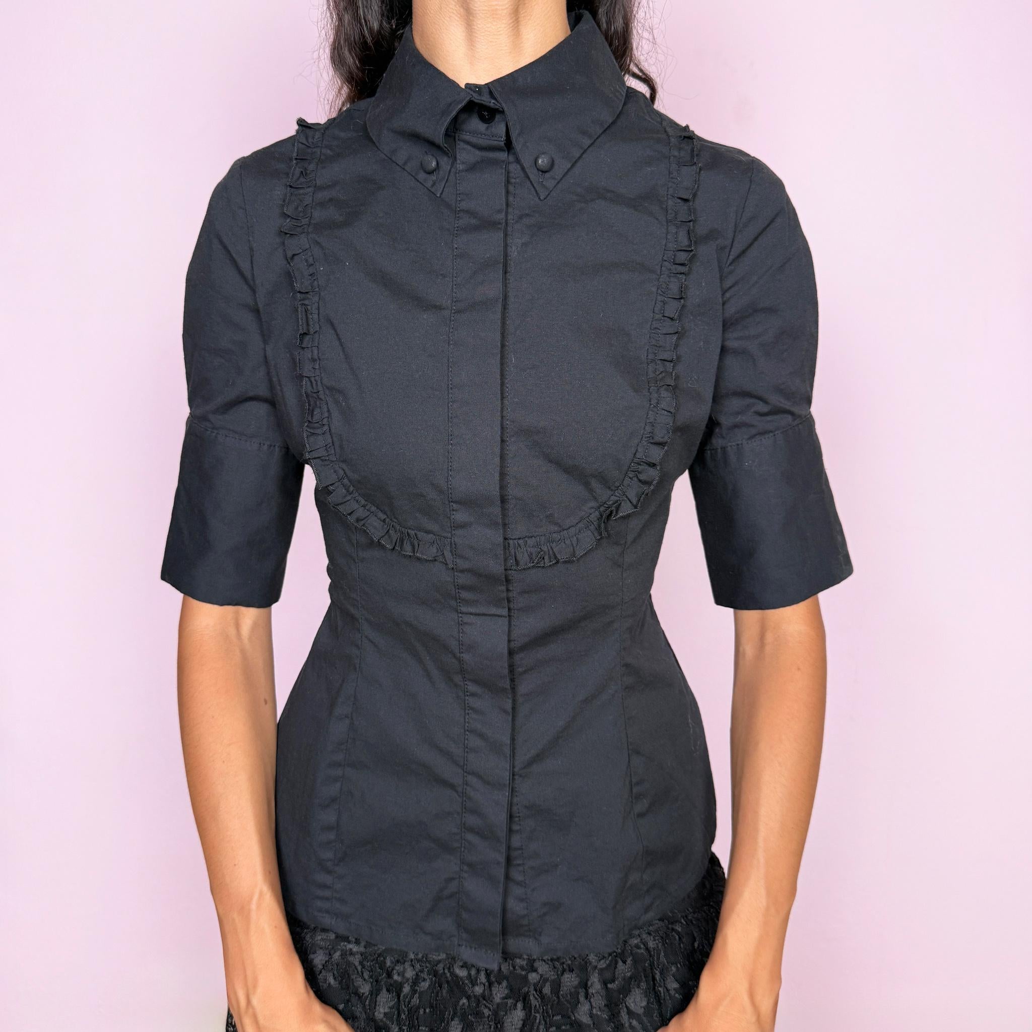 Vintage 2000s black fitted shirt top crafted from a slightly stretchy blend of 97% cotton and 3% elastane. Featuring a structured pointed collar, concealed front button closure, and sculpted panel seams that create a sharply tailored silhouette, finished with ruffled bib detailing across the chest and short sleeves. A refined piece balancing formal tailoring with subtle romantic detailing, ideal for elegant minimalist or dark feminine styling. By Spanish designer Adolfo Domínguez.