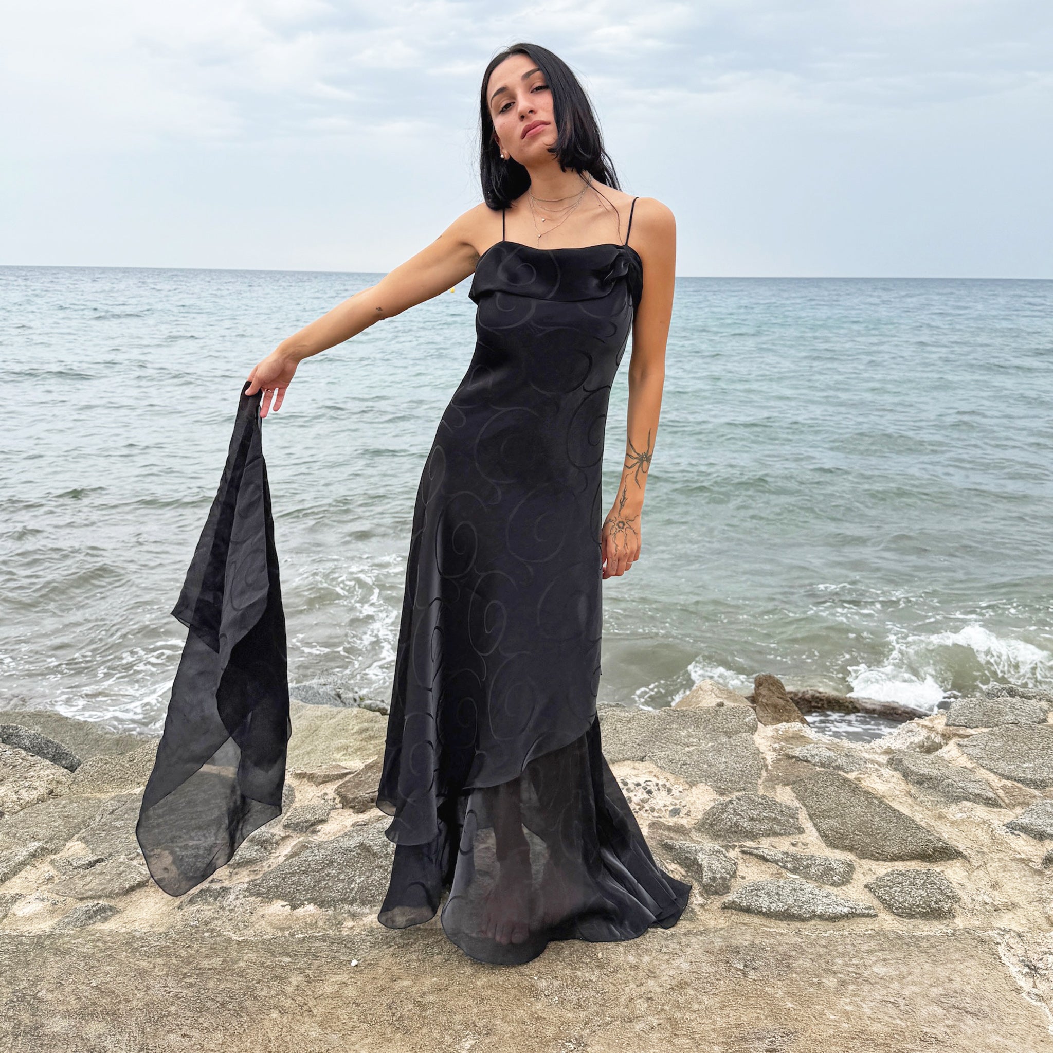 Vintage 2000s statement black maxi dress with iridescent shimmer, elegant layered construction, spaghetti straps, asymmetrical tiered hem, and sheer swirl-pattern overlay. Fluid body-skimming fit with a matching scarf, perfect for evening events, occasionwear, proms, or dark romantic nights. Made in France by a French brand from non-stretch 78% viscose and 22% polyester with 100% polyester lining. Excellent vintage condition.