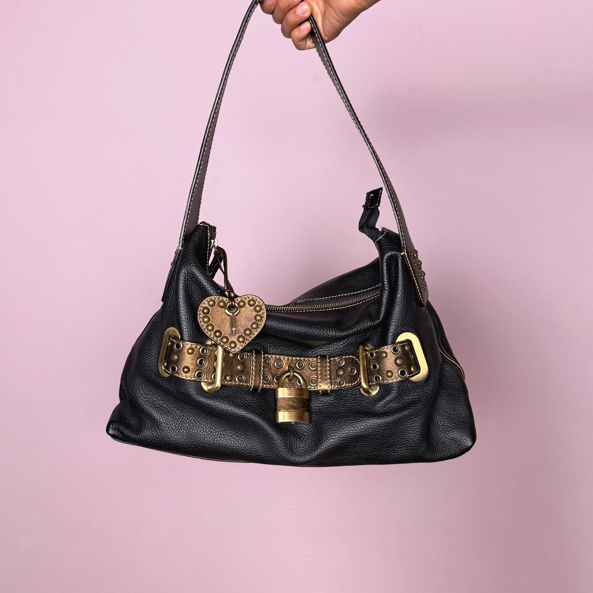 Vintage 2000s black genuine leather shoulder bag made in Italy, Firenze. Features a soft slouchy silhouette, top zip closure, bronze and gold-tone belted front detail, metal eyelets, studded hardware, heart charm, and decorative padlock accent. A unique Y2K Italian leather statement bag with a biker-boho glam and indie grunge feel.