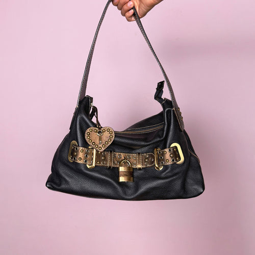 Vintage 2000s black genuine leather shoulder bag made in Italy, Firenze. Features a soft slouchy silhouette, top zip closure, bronze and gold-tone belted front detail, metal eyelets, studded hardware, heart charm, and decorative padlock accent. A unique Y2K Italian leather statement bag with a biker-boho glam and indie grunge feel.