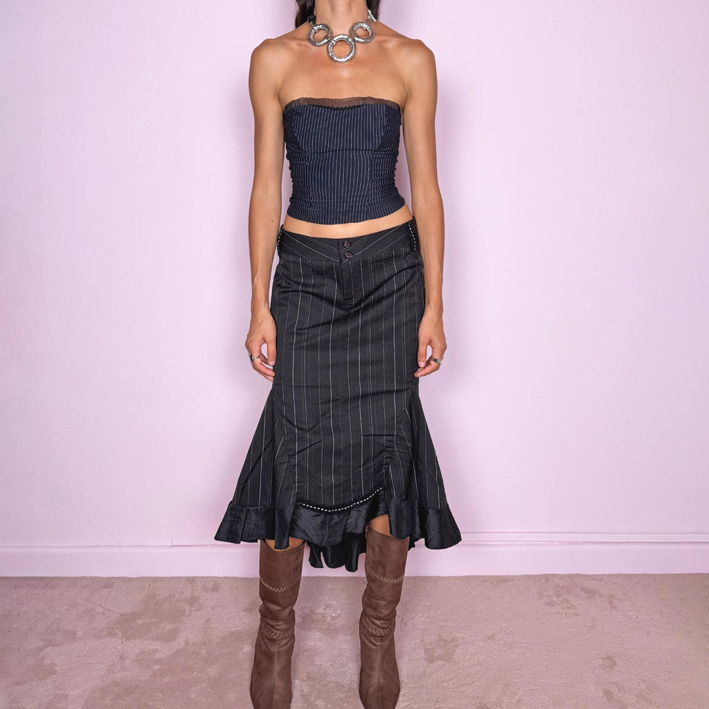 Vintage 2000s black pinstripe midi skirt featuring a fitted low-rise silhouette with a diagonal waistband construction, front zip closure, vertical pinstripes, and sculptural paneling through the lower skirt. Finished with an asymmetric flared hem and contrasting satin-like ruffle trim for a dramatic, deconstructed Y2K silhouette. Made in Italy by an Italian brand from a slightly stretchy blend of 62% polyester, 33% viscose, and 5% elastane. Excellent vintage condition.