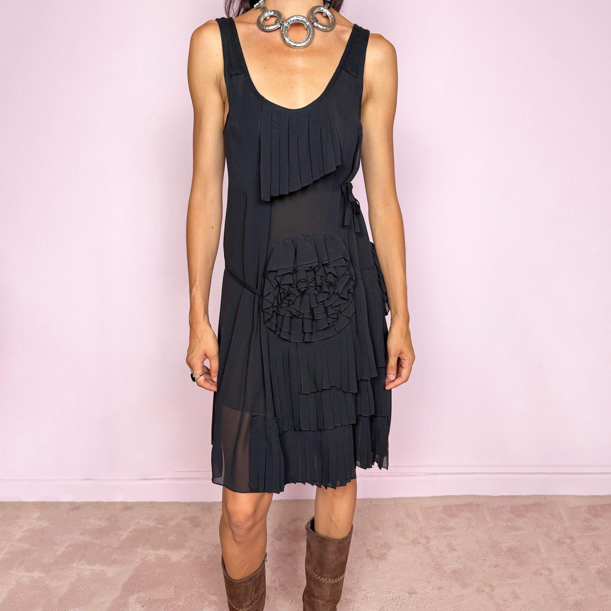 Vintage 2000s black midi dress with a soft sheer layered finish. It features a sleeveless scoop neckline, relaxed shift silhouette, side tie details, and an asymmetric overlay decorated with sculptural pleated appliqués, rosette-style fabric detailing, and layered chiffon panels that add depth and movement. A refined Italian piece with an avant-garde, deconstructed art-to-wear feel, ideal for a minimal elegant evening look. Made by Patrizia Pepe from 100% silk