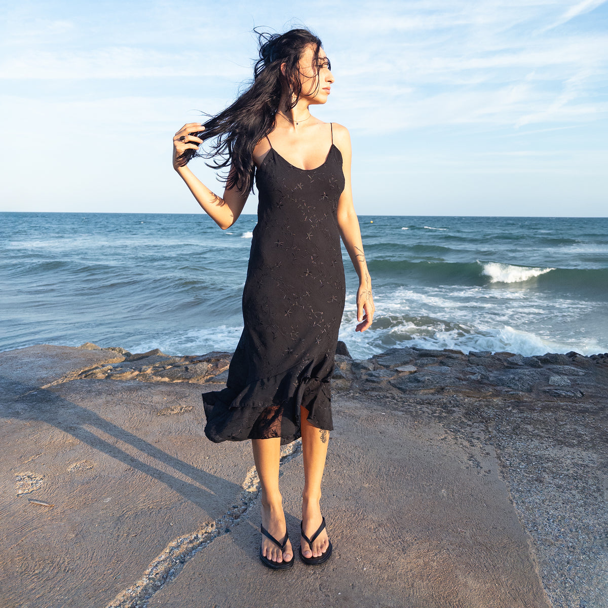 Vintage 2000s stretchy black slip midi dress featuring slim spaghetti straps, a softly fitted silhouette, subtle floral embroidery over a semi-sheer overlay with black lining underneath, and a layered ruffle hem with an asymmetric drape. Perfect for dark feminine summer evening outfits. From a French brand, made in France from 100% polyester. Excellent vintage condition.
