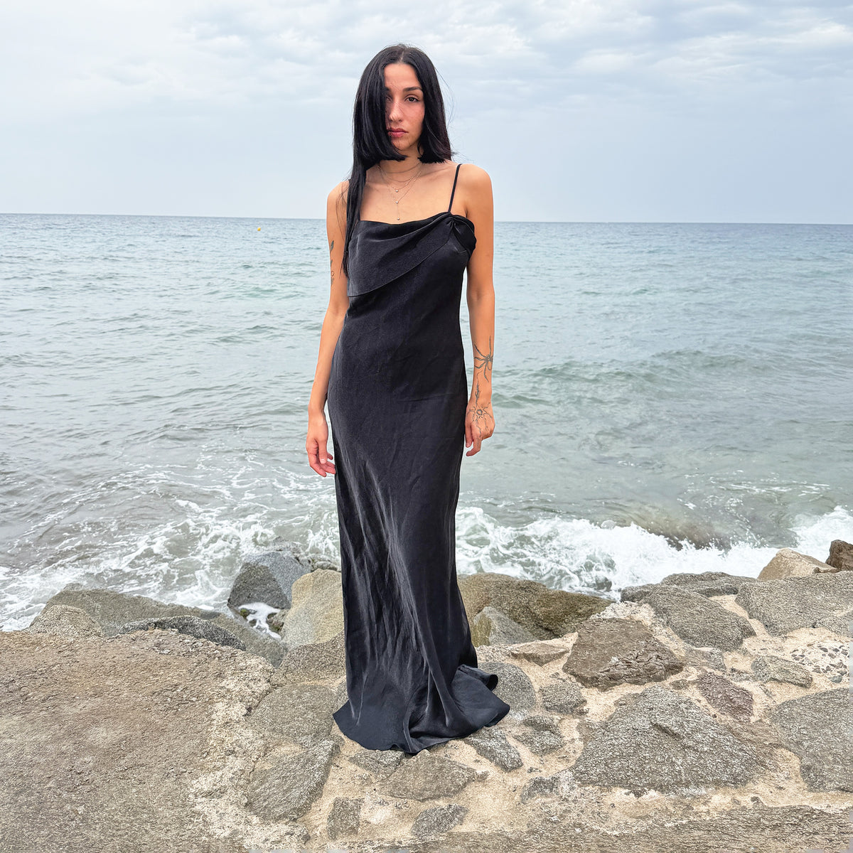 Vintage 90s black slip maxi dress featuring a sleek, body-skimming silhouette, slim spaghetti straps, and a sculptural asymmetric draped neckline with gathered detailing at one side. Cut from a non-stretch fabric with a soft satin-like sheen and finished with a side zip closure. Perfect for minimal evening styling and dark romantic looks. Composition label is missing, so the exact fabric is unknown. Excellent vintage condition.