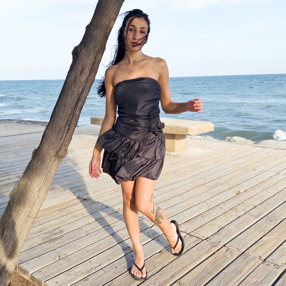 Vintage 90s black strapless mini dress made in Italy, featuring a sculptural fitted bodice with gathered ruching, a dropped waist accented with a fabric rosette, and a voluminous double-tiered ruffle skirt. Designed with a shirred elastic back panel, tie-back detail, and side zip closure. Perfect for cocktail styling, summer evening looks, and playful dark feminine outfits. Made from a blend of 95% polyester and 5% elastane. Excellent vintage condition.