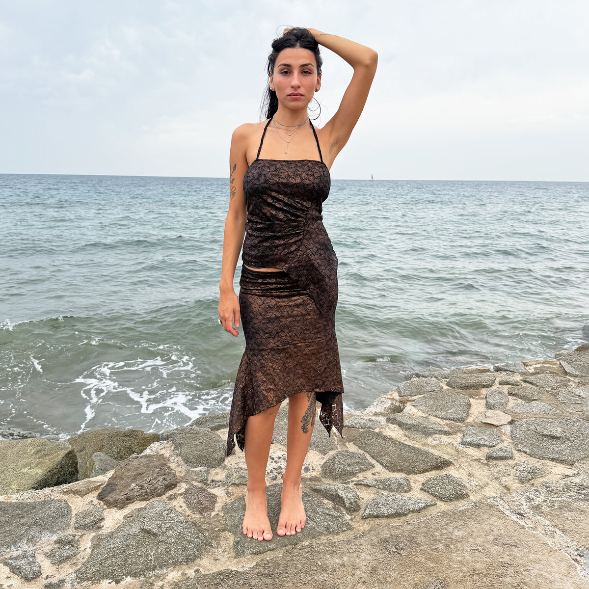 Vintage 2000s two-piece set in a deep bronze-brown tone, made in Spain by a Spanish designer. Crafted from stretchy black floral lace with a metallic bronze-copper coated finish. The set includes a fitted halter top with ruched detailing, tie-neck fastening and an asymmetric pointed hem, paired with a matching handkerchief-style midi skirt featuring an elastic waistband. The skirt can be worn mid-rise or low-rise on sizes XS and S. Perfect for dark summer evening looks and festival outfits. Made from a stre
