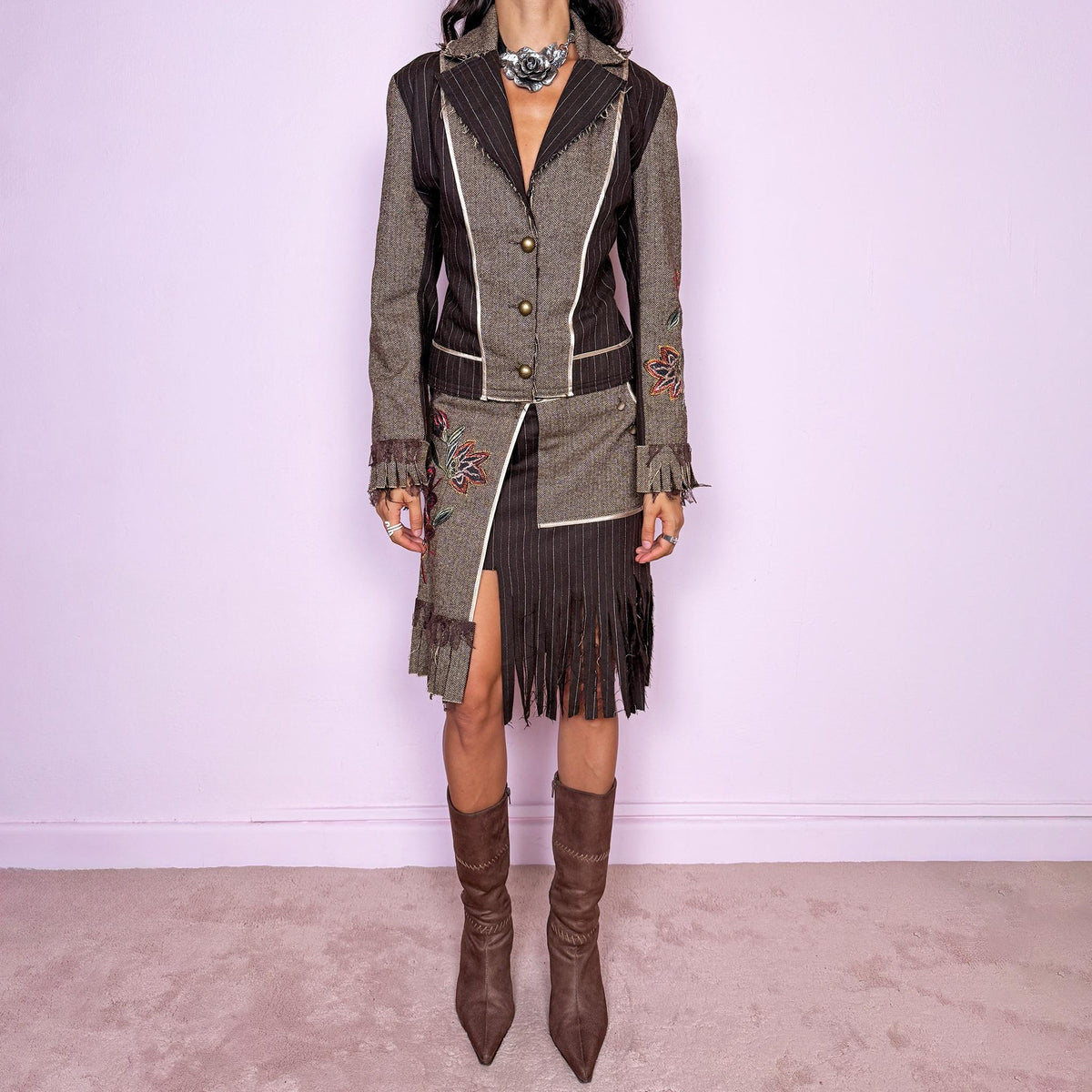 Vintage 2000s deconstructed two-piece skirt suit. The fitted jacket features mixed pinstripe and herringbone panels, raw-edge detailing, contrast piping, floral embroidery, decorative metal buttons, and pleated fringe cuffs. Paired with a matching asymmetric midi skirt featuring layered panel construction, a side zip closure, embroidered floral motifs, and dramatic cut fringe through the lower section. Non-stretch.