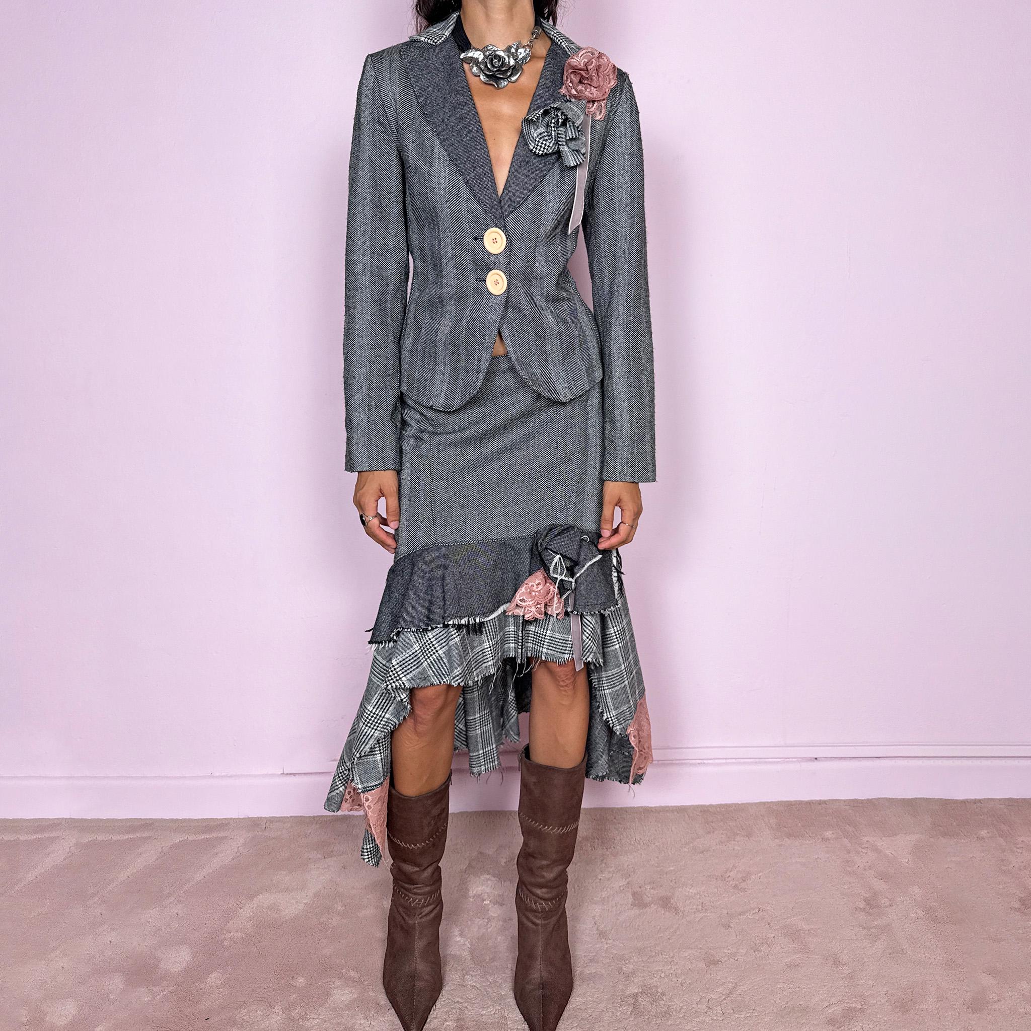 Vintage 2000s two-piece skirt suit crafted from an 80% wool and 20% polyamide blend with no stretch. The fitted gray herringbone blazer features contrasting oversized buttons, wide lapels, and sculptural fabric flower embellishments in dusty pink and monochrome checks, while the matching mid-rise midi skirt combines multiple herringbone and plaid panels with distressed raw edges, asymmetric cascading ruffles, romantic floral appliqué details, and a side zip closure.