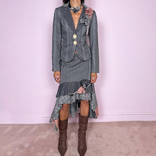 Vintage 2000s two-piece skirt suit crafted from an 80% wool and 20% polyamide blend with no stretch. The fitted gray herringbone blazer features contrasting oversized buttons, wide lapels, and sculptural fabric flower embellishments in dusty pink and monochrome checks, while the matching mid-rise midi skirt combines multiple herringbone and plaid panels with distressed raw edges, asymmetric cascading ruffles, romantic floral appliqué details, and a side zip closure.