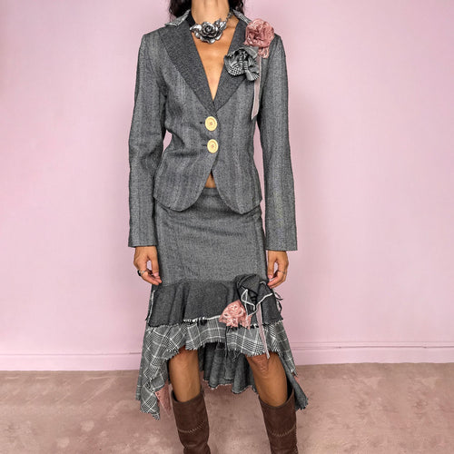 Vintage 2000s two-piece skirt suit crafted from an 80% wool and 20% polyamide blend with no stretch. The fitted gray herringbone blazer features contrasting oversized buttons, wide lapels, and sculptural fabric flower embellishments in dusty pink and monochrome checks, while the matching mid-rise midi skirt combines multiple herringbone and plaid panels with distressed raw edges, asymmetric cascading ruffles, romantic floral appliqué details, and a side zip closure.