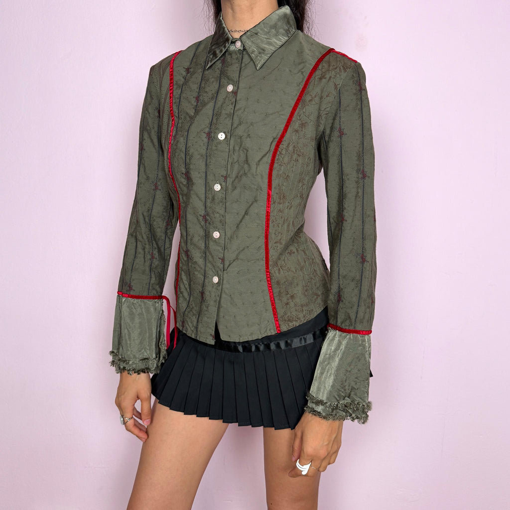 Vintage 2000s muted olive green blouse with subtle floral texture, button-front closure, sharp pointed collar, vertical paneling, contrasting red velvet trim, satin-like collar and cuff details, flared sleeves, and softly frayed edges for elevated earthy styling. Composition label is missing, so the exact fabric is unknown. Great vintage condition.