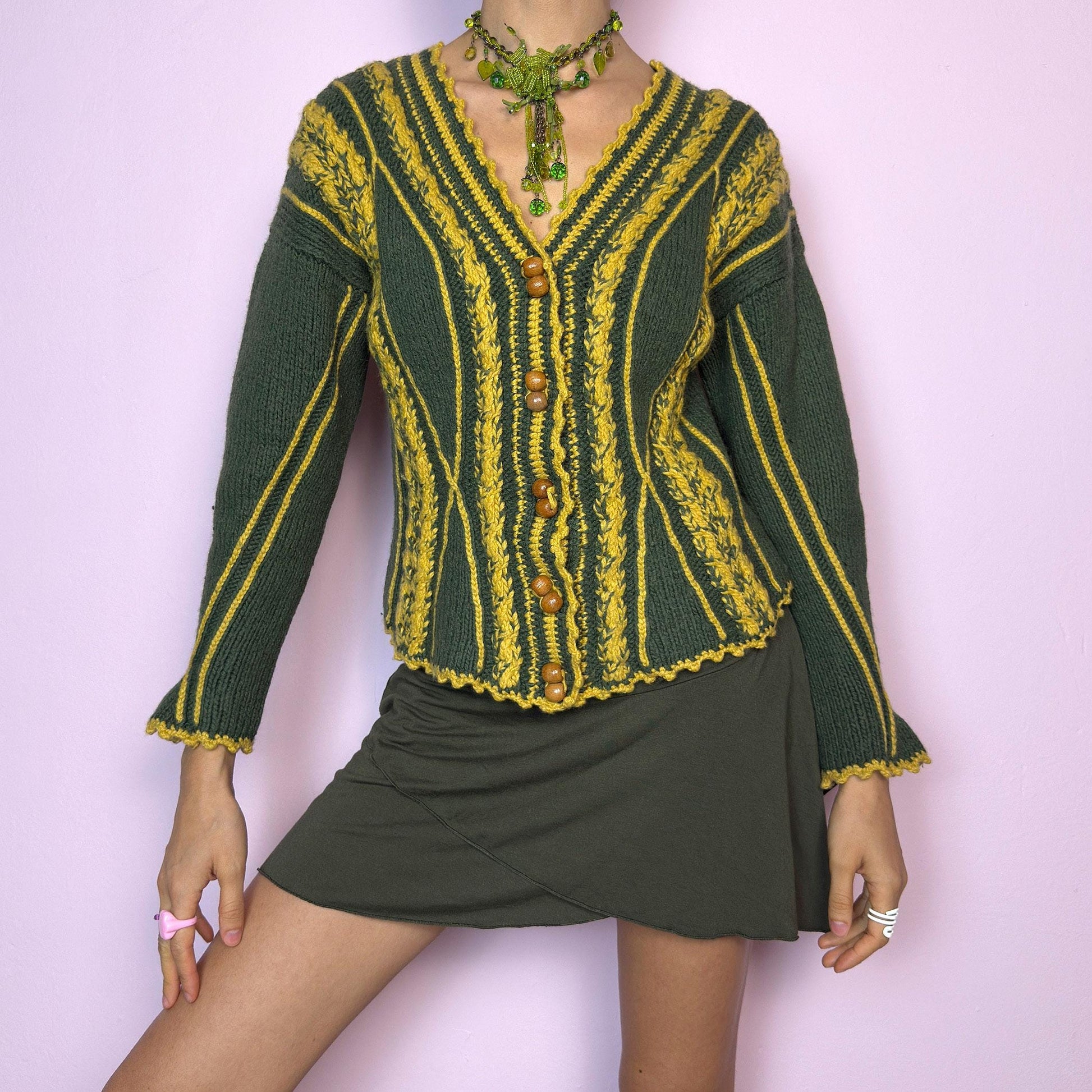 The Vintage Handmade-Style Chunky Knit Cardigan features intricate cable knit detailing in green and mustard yellow. With its flared cuffs, scalloped hems, and wooden buttons, this piece evokes a folk-inspired or alpine feel, reminiscent of 70s European ski towns and handmade market finds. The fitted silhouette and vertical lines create a flattering shape while adding bold texture. A one-of-a-kind layering piece perfect for forest walks, cozy days, or cottagecore styling.  adult world shop